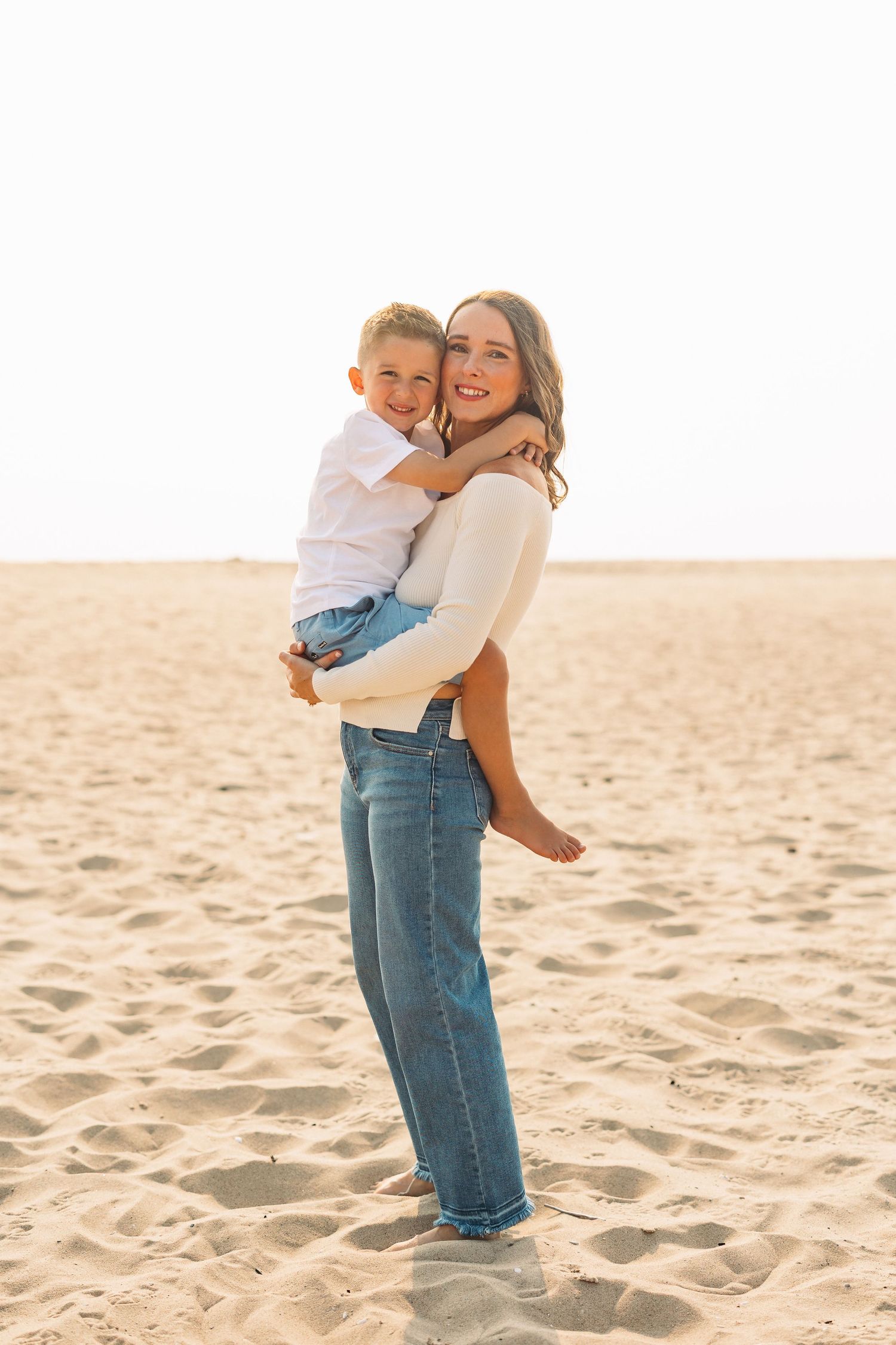 Smiling mother holding young son on a sunny sandy beach, both wearing white tops and jeans.