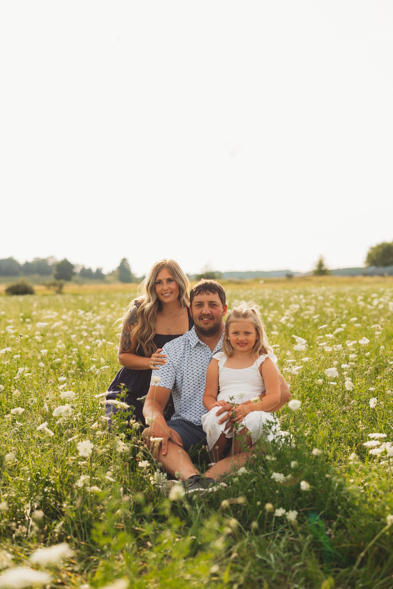 Family of three sitting in a sunny wildflower field, parents smiling with young daughter in white dress.