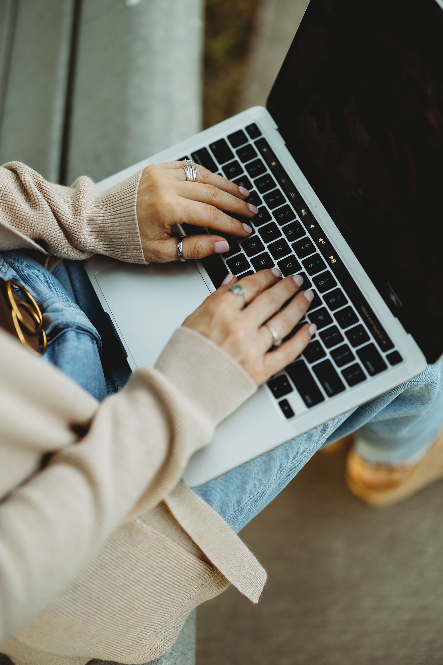 Close-up of woman typing on a silver laptop while wearing a beige cardigan and rings.