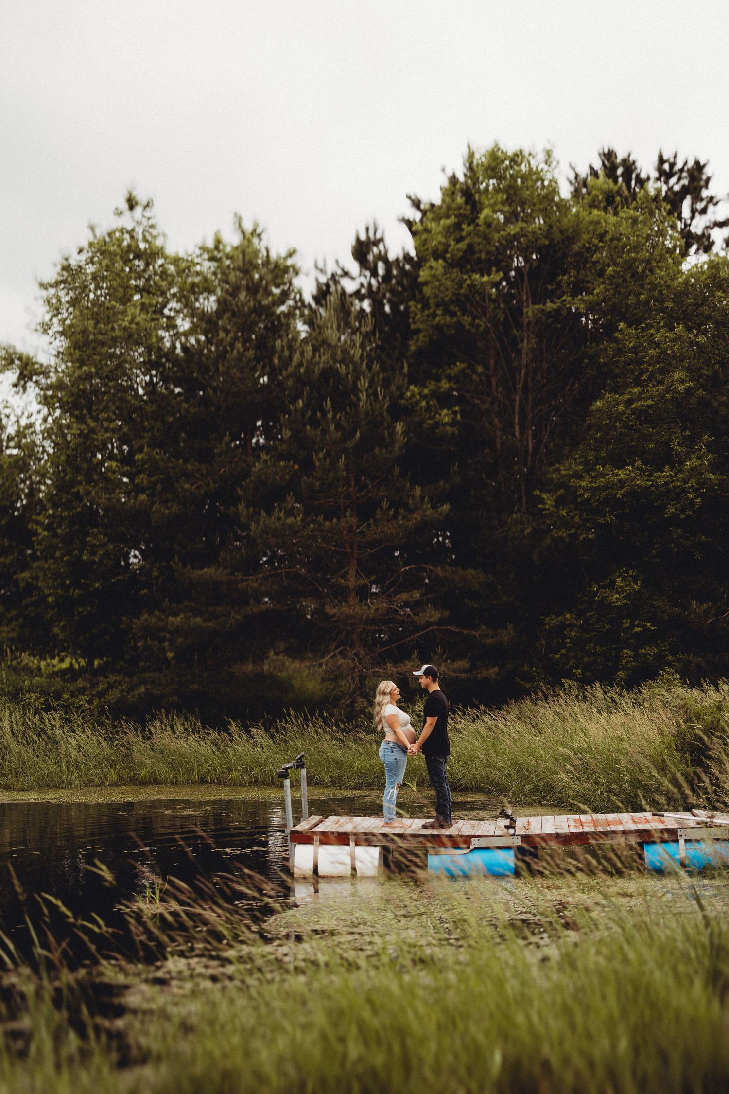 Couple standing on a rustic dock by a serene pond surrounded by lush green trees and tall grass.