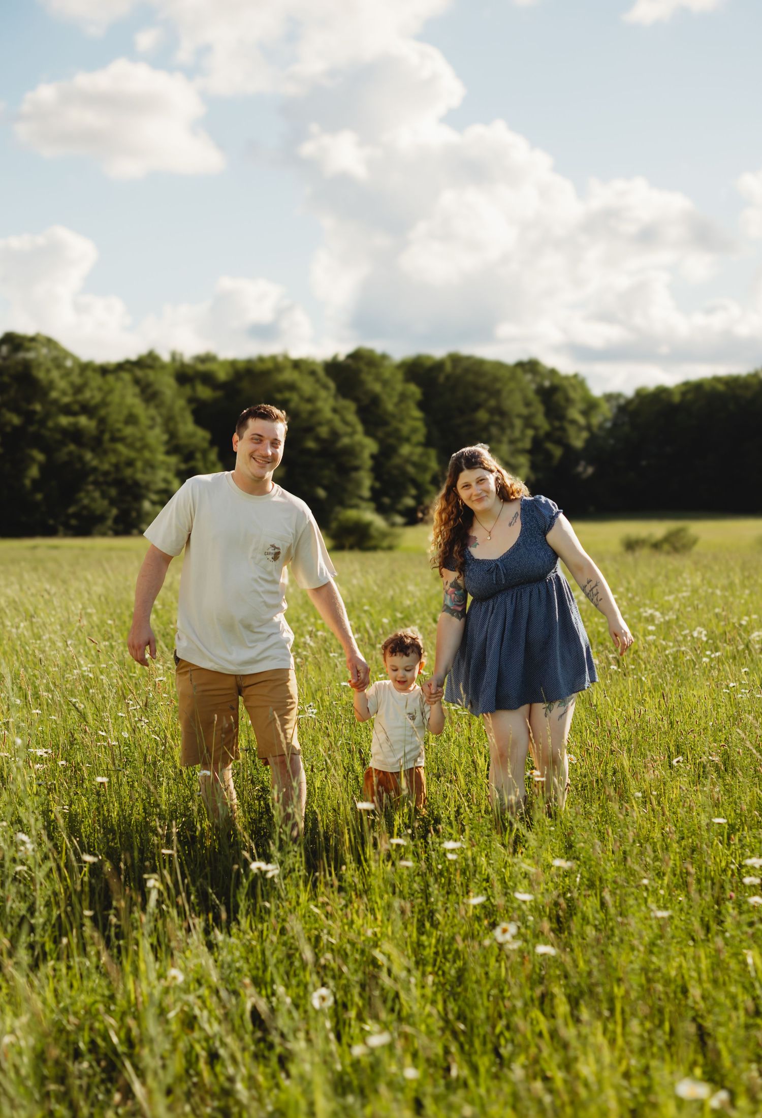A family of three walks hand-in-hand through a sunny green meadow with wildflowers, trees, and blue sky behind them.