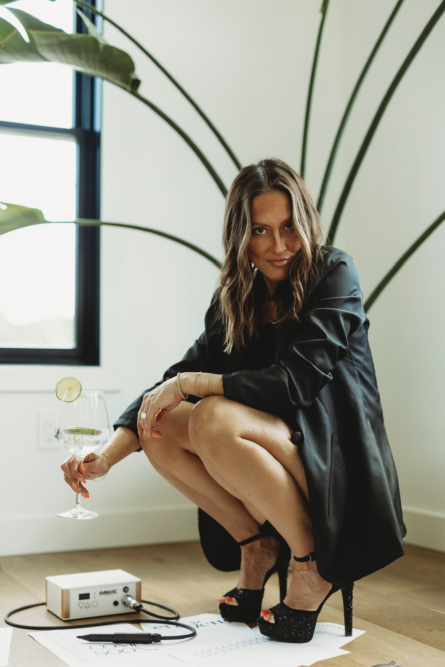 Woman in black dress holding wine glass, crouching near plant and electronic device on wooden floor.
