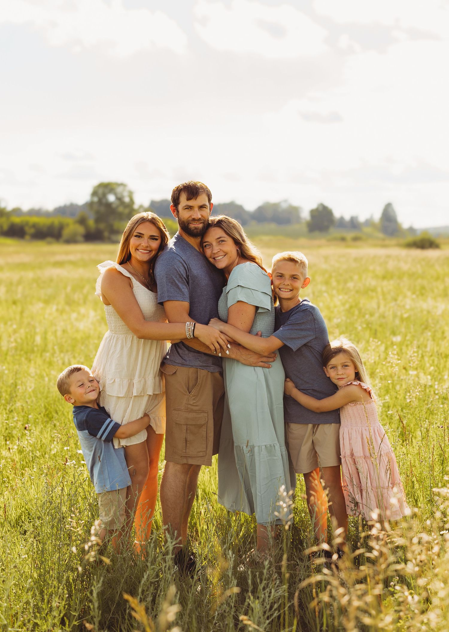 Family of six posing together in a sunny green meadow, smiling warmly in coordinated casual outfits during golden hour.
