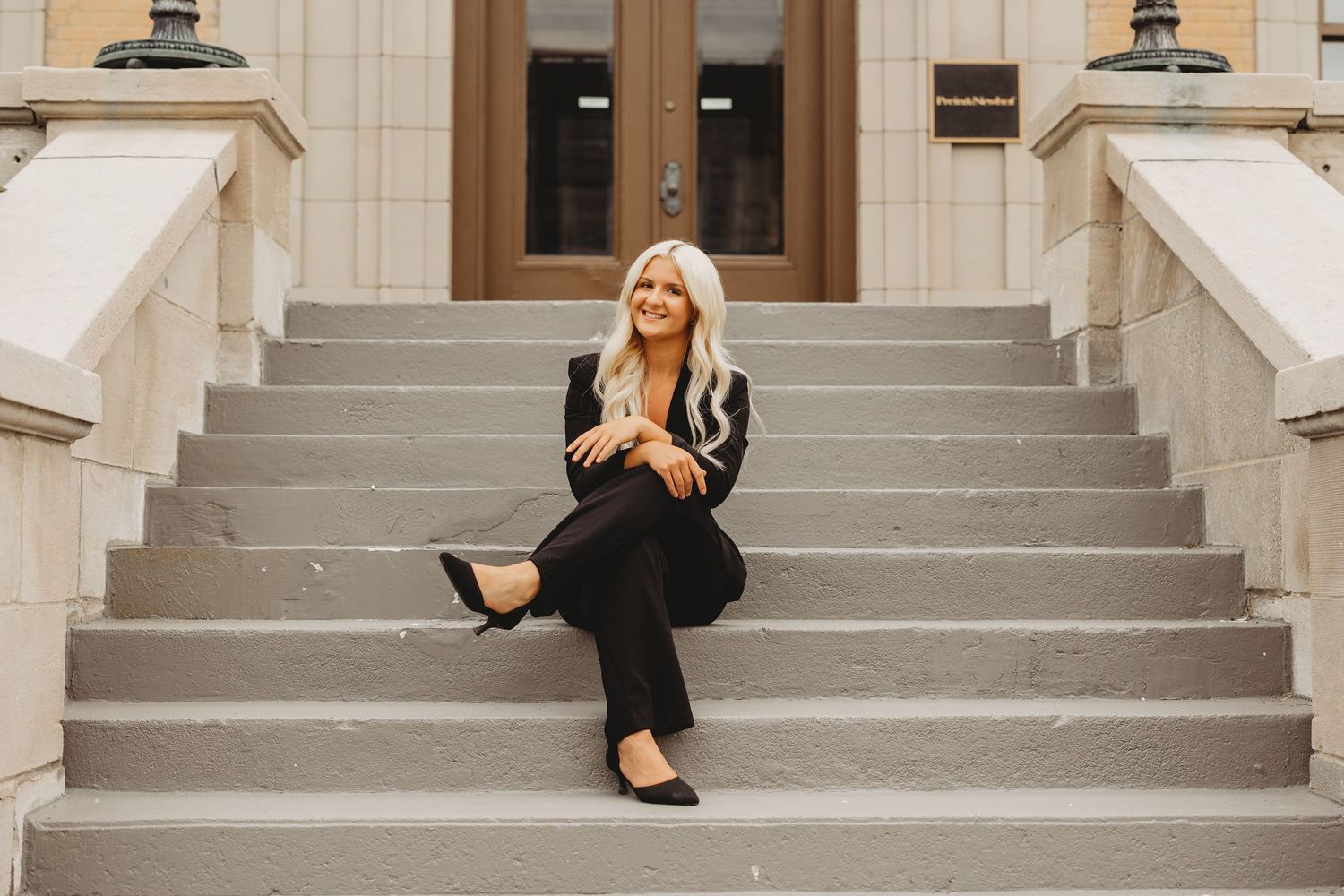 A smiling blonde woman in a black outfit sits on gray stone steps outside a classic building.