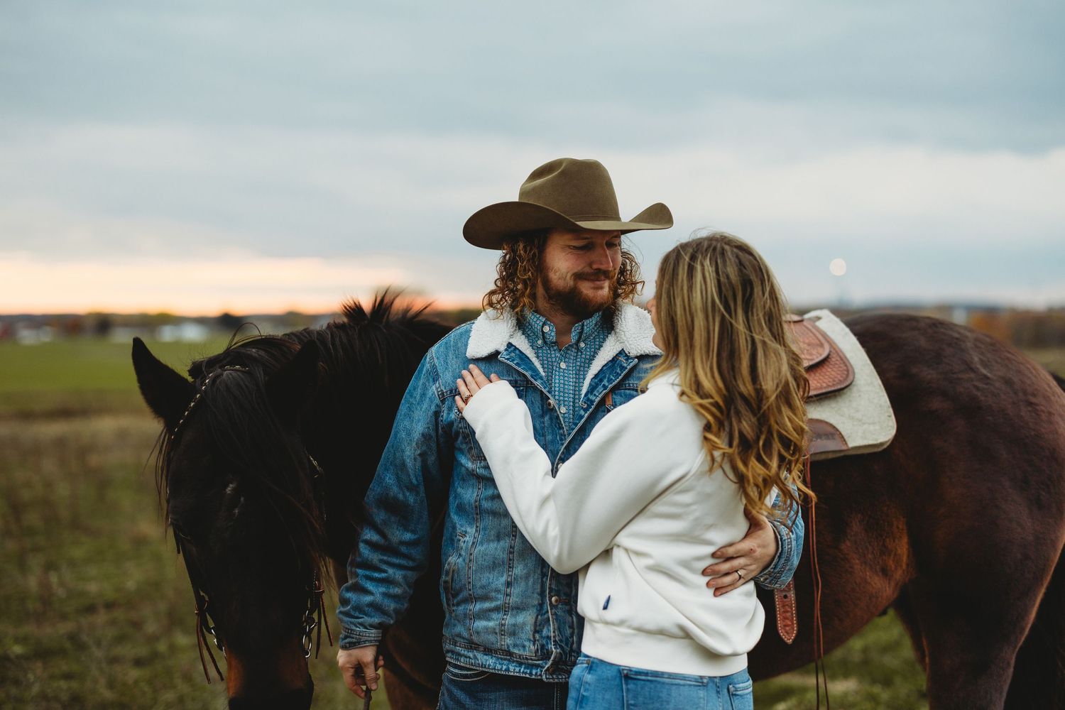 A couple in western attire embraces in a golden field at sunset with horses in the background.
