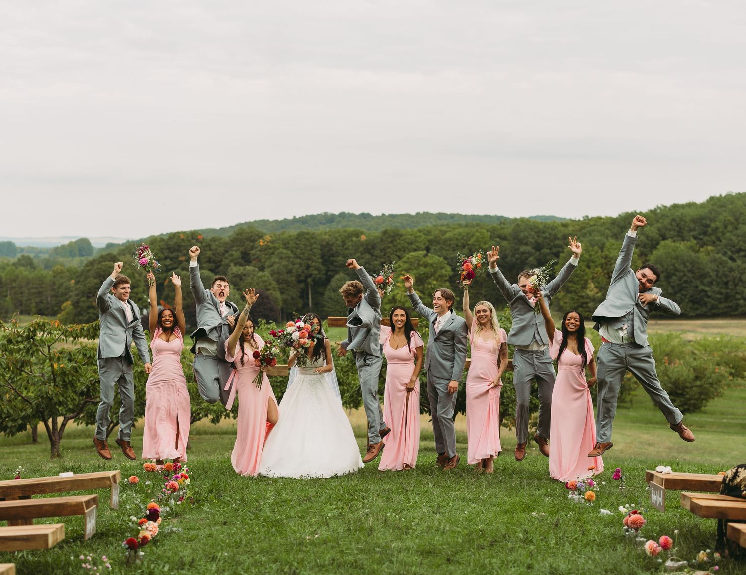 Large wedding party celebrates on a hillside with bridesmaids in pink and groomsmen in gray suits.