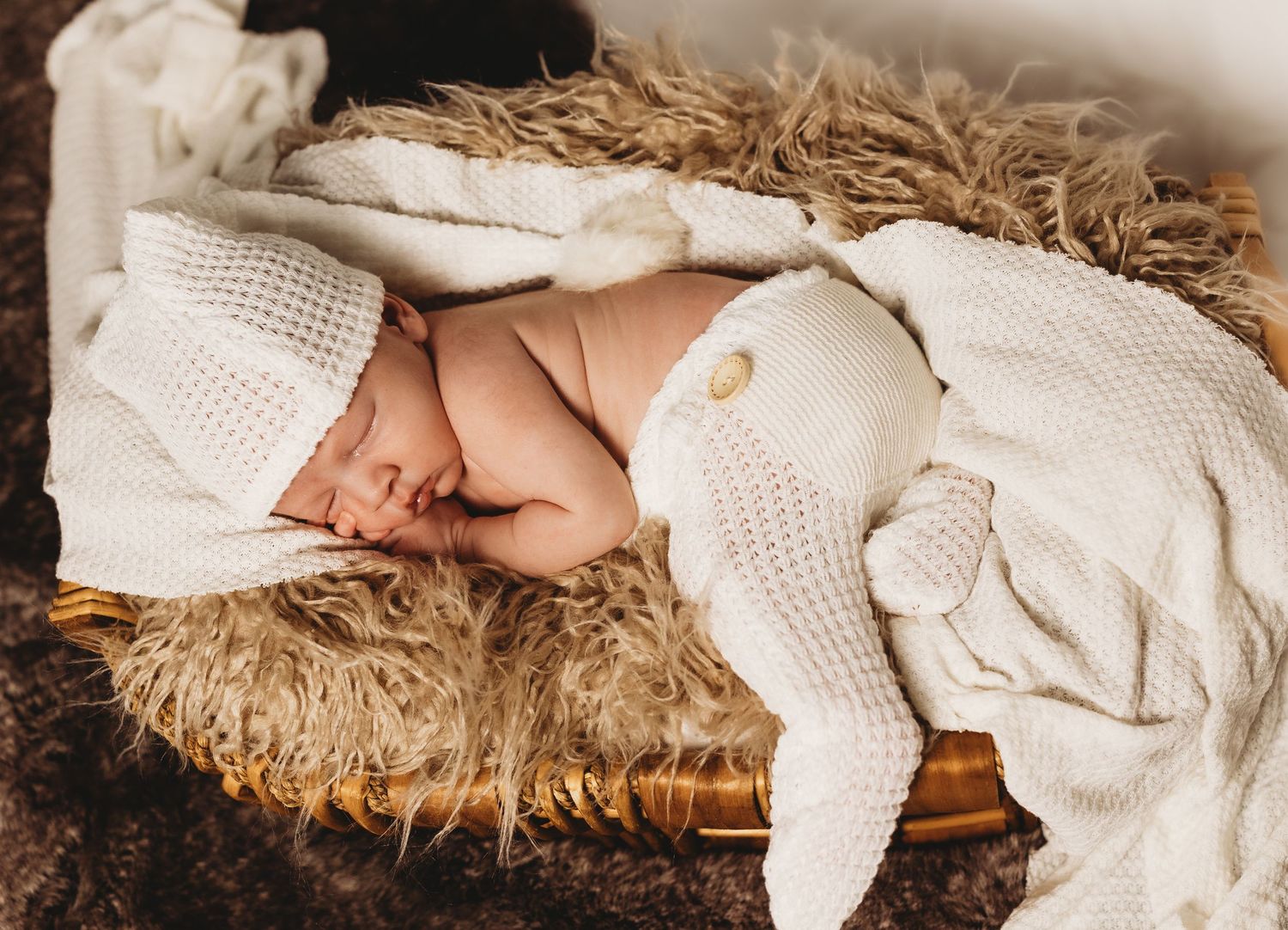 Newborn baby wrapped in white blanket resting on soft fur blanket with Christmas lights in background.