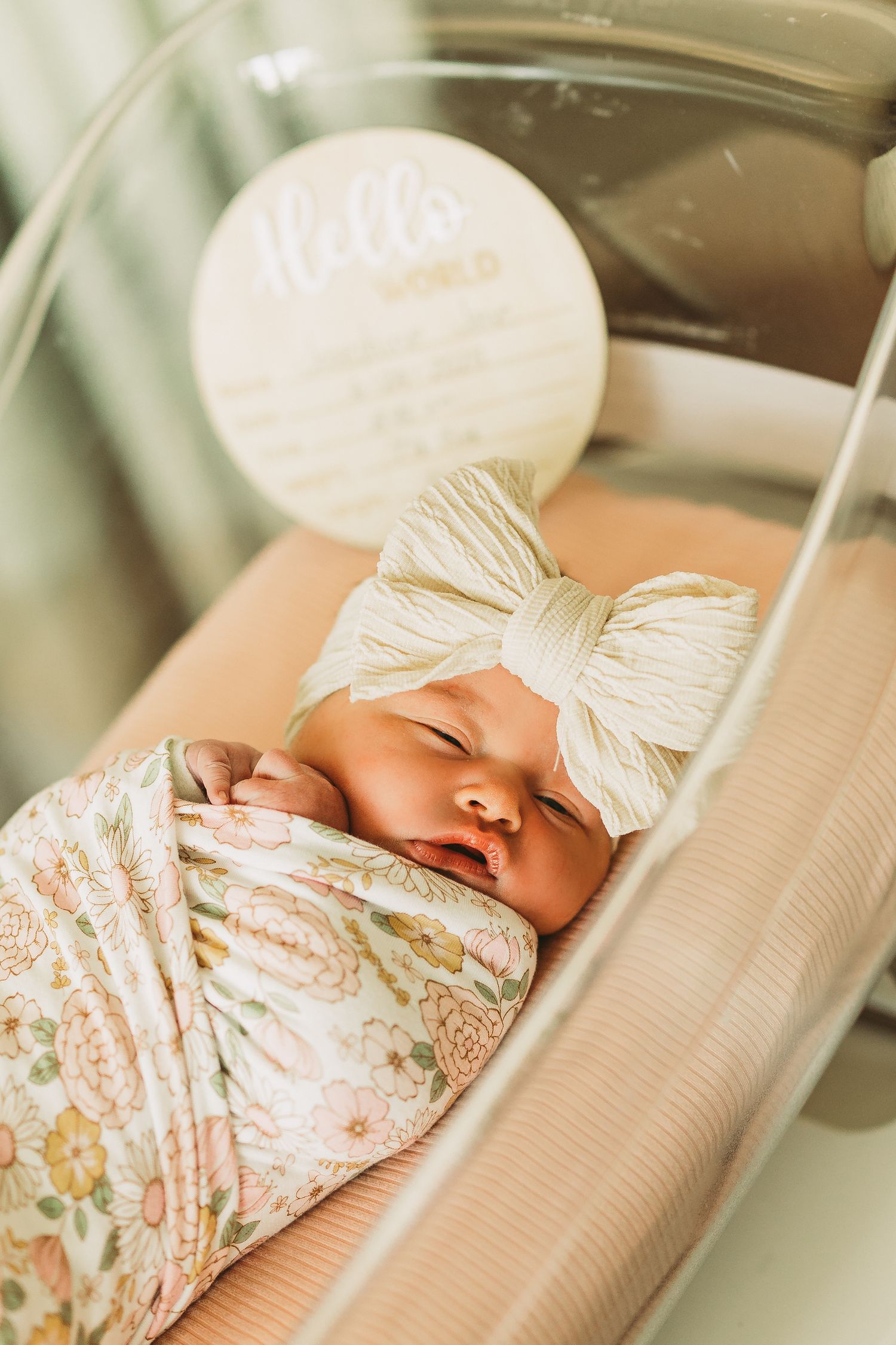 Sleeping newborn wrapped in floral swaddle blanket with white bow headband.
