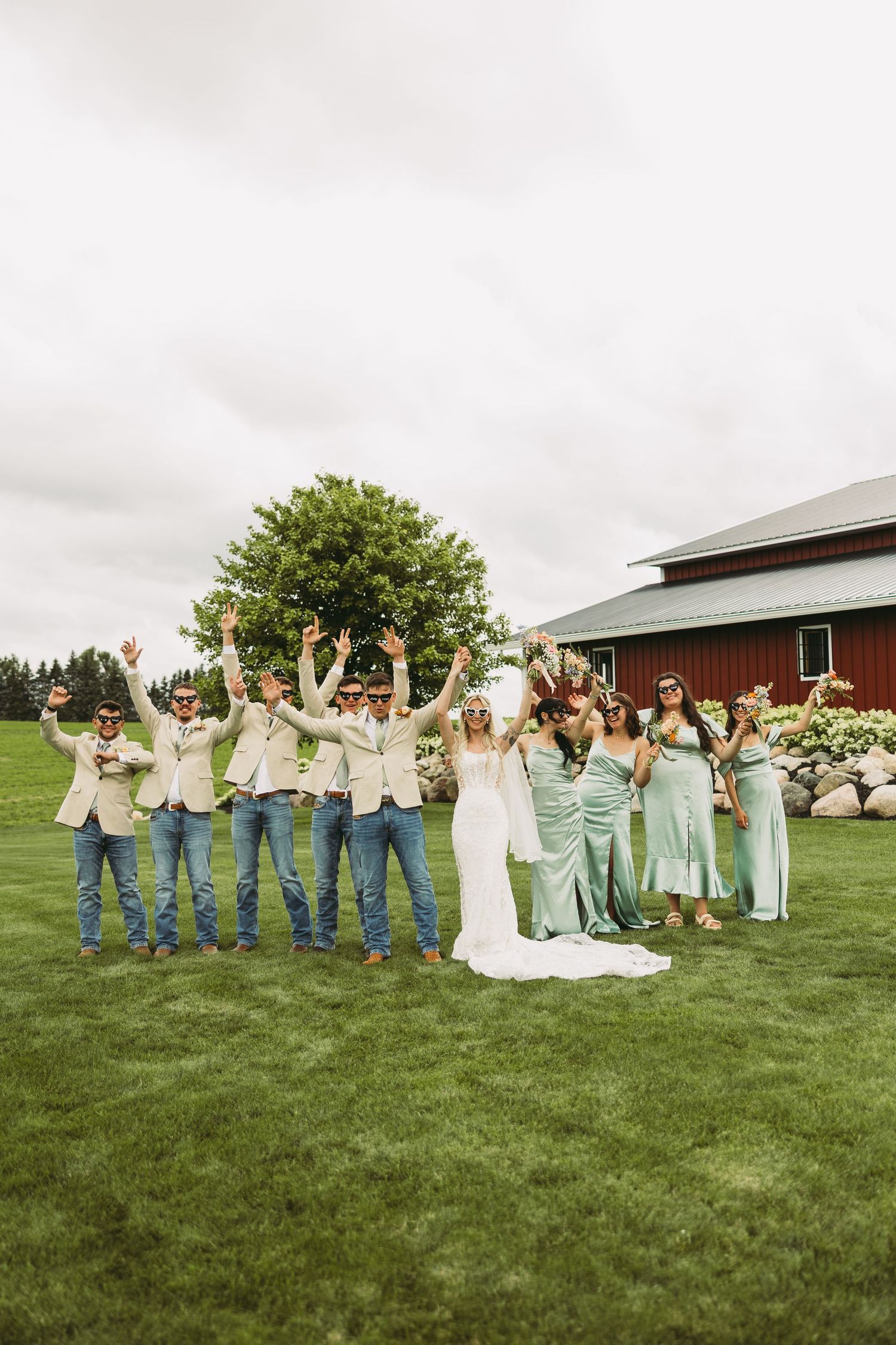 Wedding party in jeans and light colored tops poses joyfully on green grass with red barn in background.