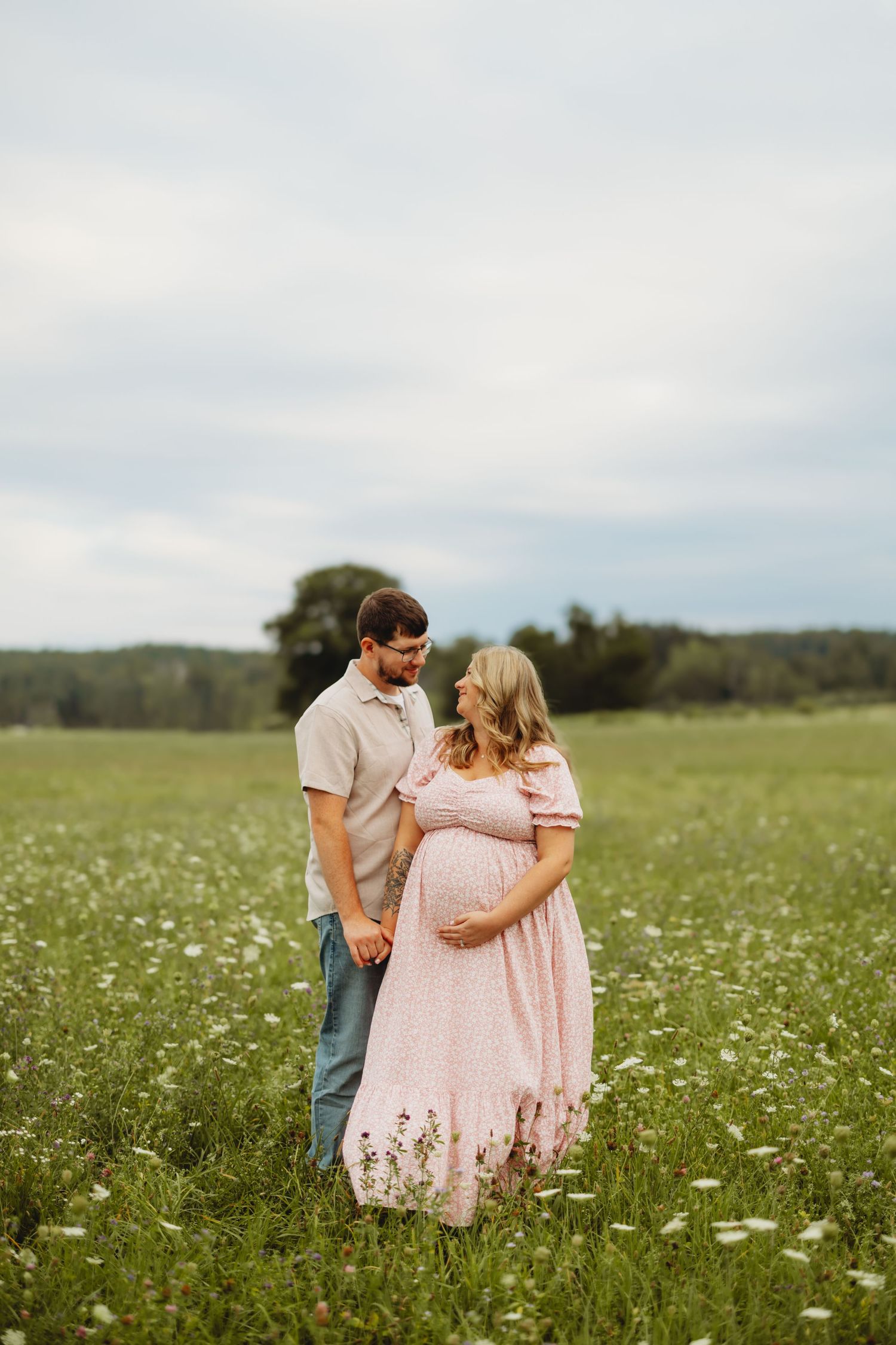 Couple sharing a tender moment in a blooming meadow, pregnant woman in pink floral dress holding hands.
