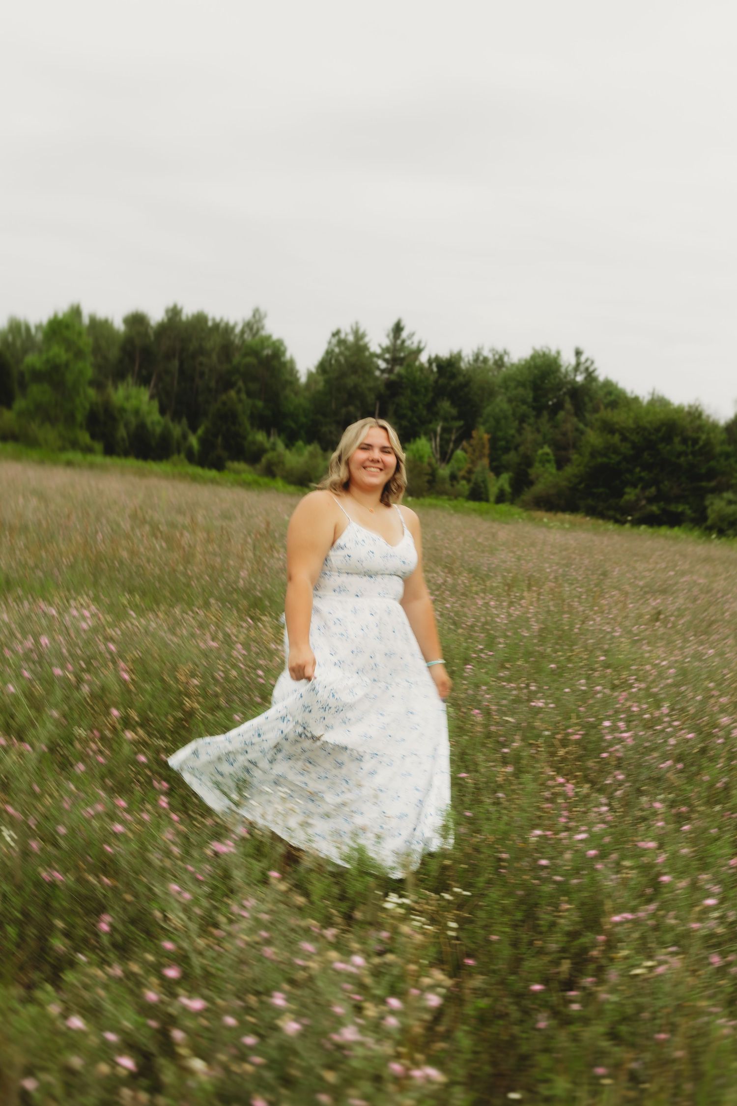 Smiling woman in white floral sundress twirling in a wildflower meadow with green trees in the background.