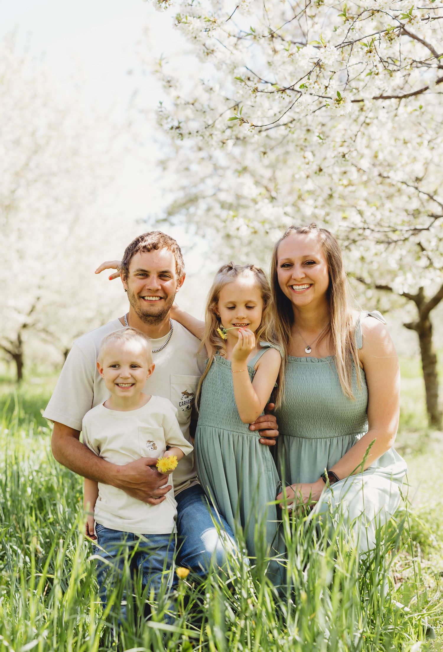 A happy family of four poses together in a blooming orchard with white flowering trees and green grass.