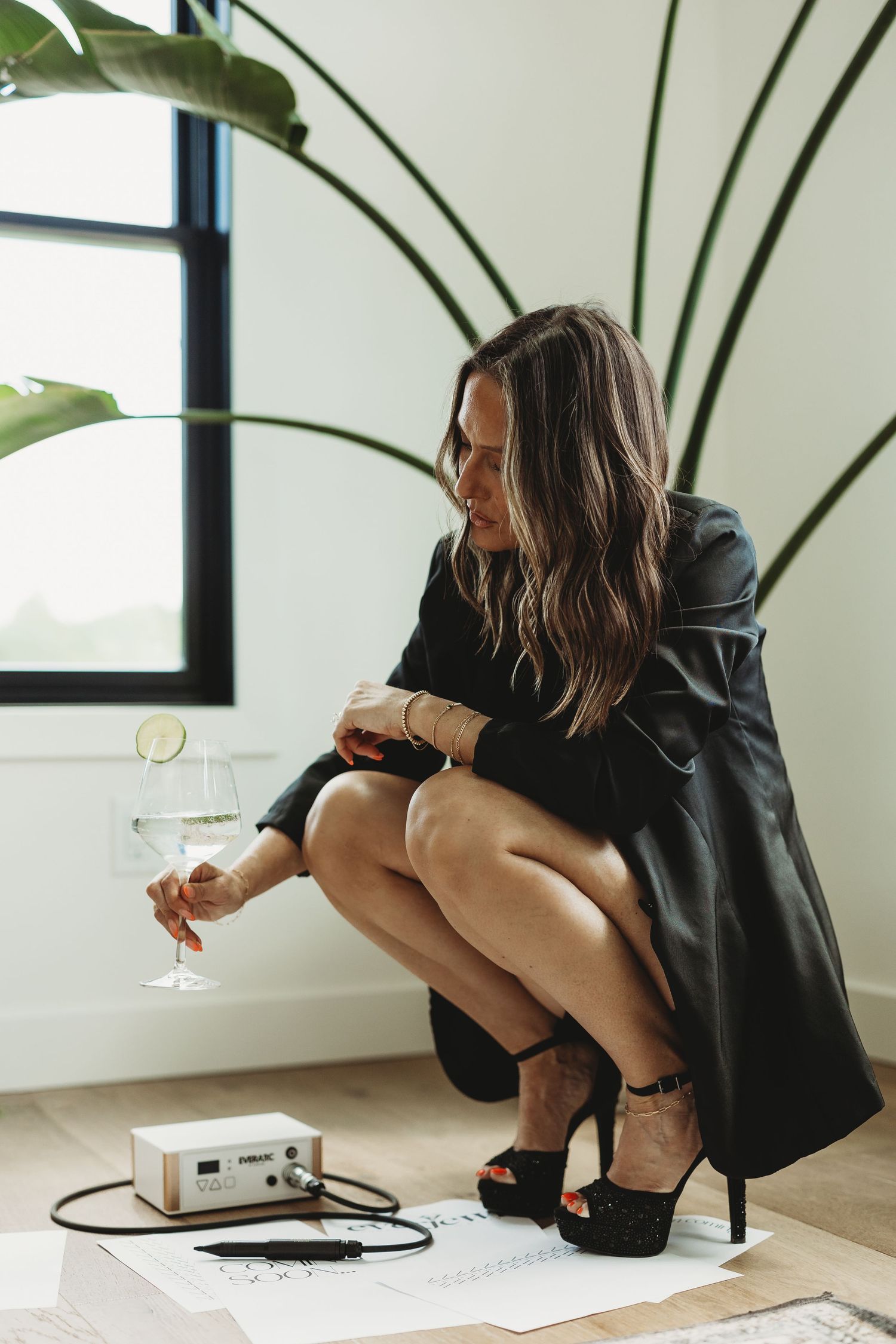 Woman in black leather dress holding wine glass, crouching near audio equipment on wooden floor with tropical plant backdrop.