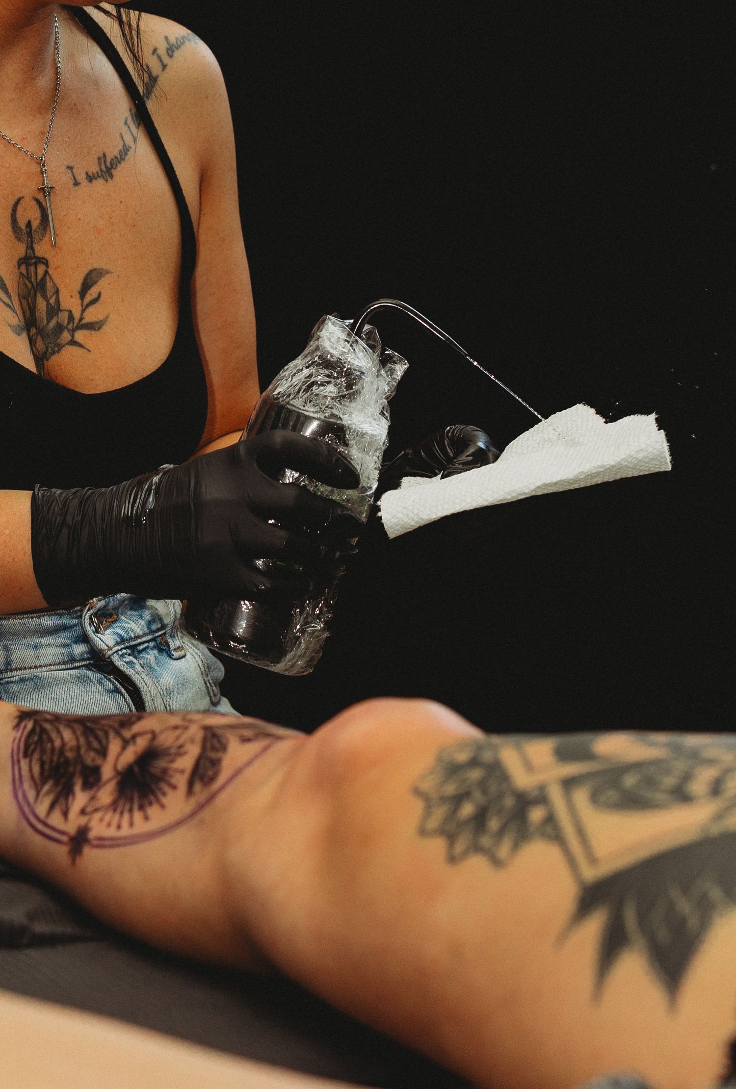 Tattooed female tattoo artist wearing black gloves cleaning tattoo equipment with paper towel during session.