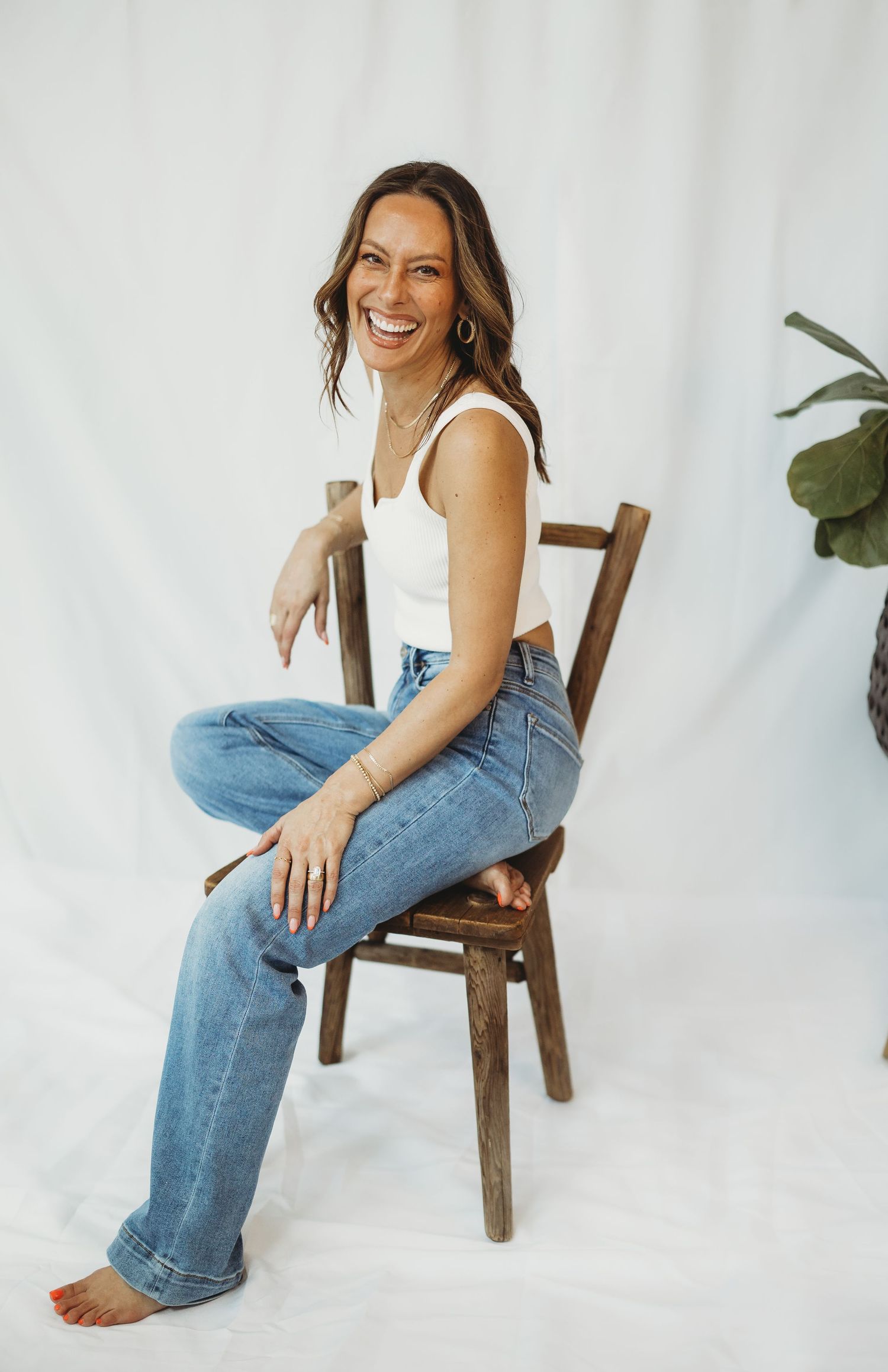 A smiling woman sits cross-legged on a wooden chair, wearing a white tank top and blue jeans, against a white backdrop.