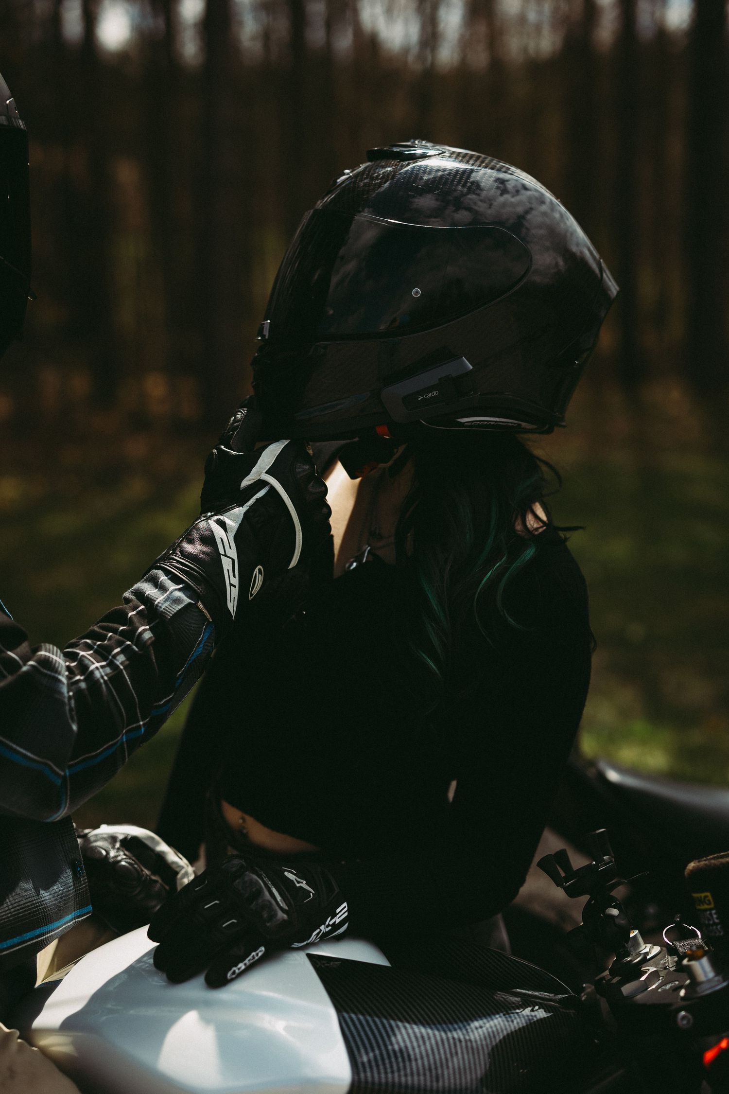 Motorcyclist in black helmet and leather gear sitting on a bike in a forest setting.