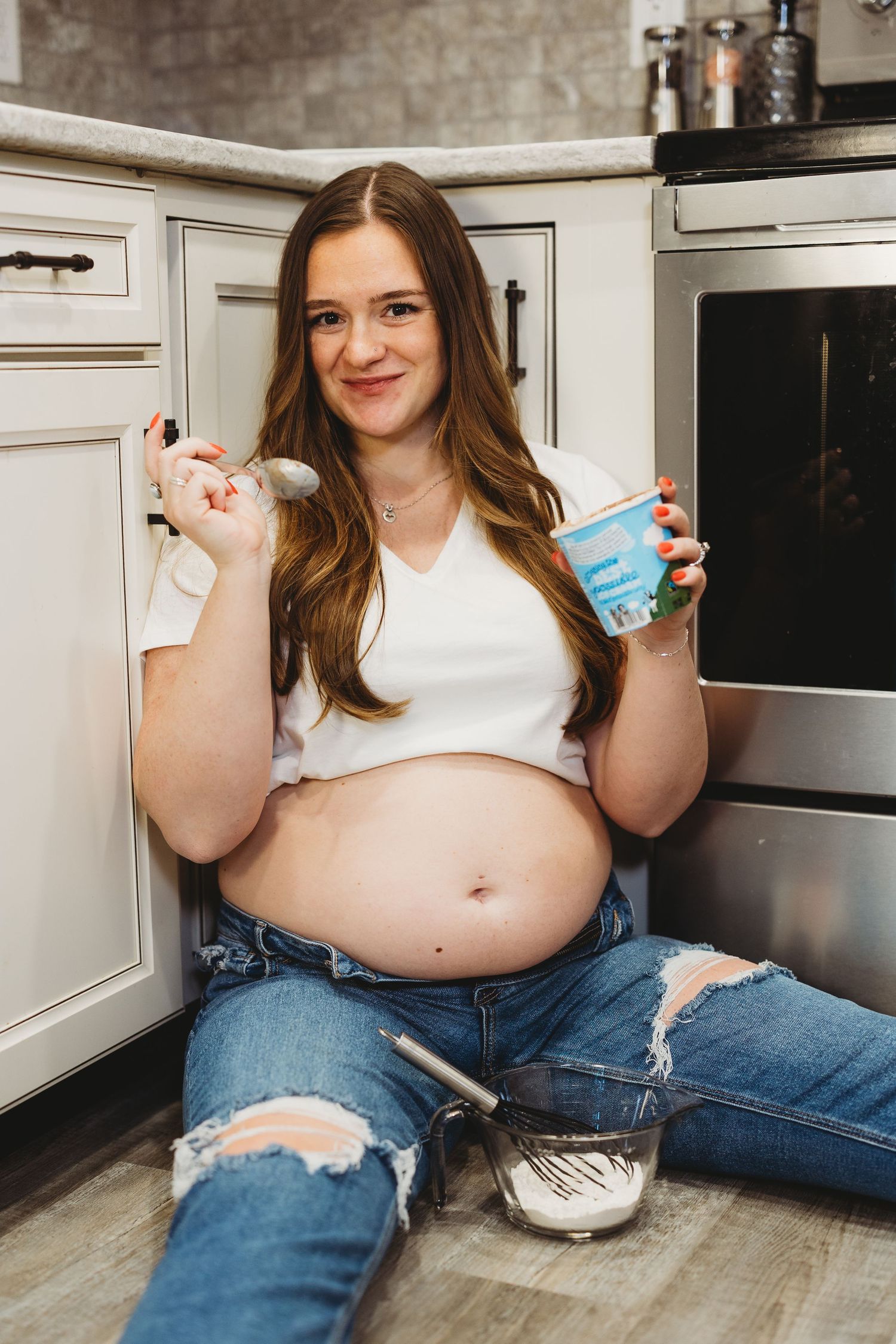 Pregnant woman sitting on kitchen floor eating ice cream with a mixing bowl nearby, wearing white crop top and ripped jeans.