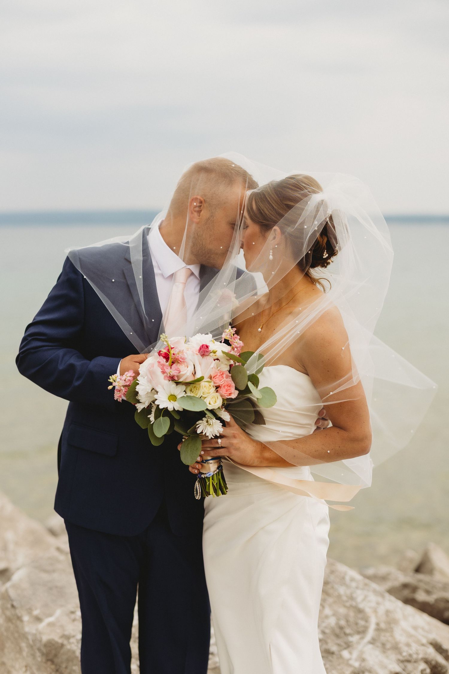Bride and groom share a tender kiss under a flowing veil by the rocky seaside, bride holding a pink and white bouquet.
