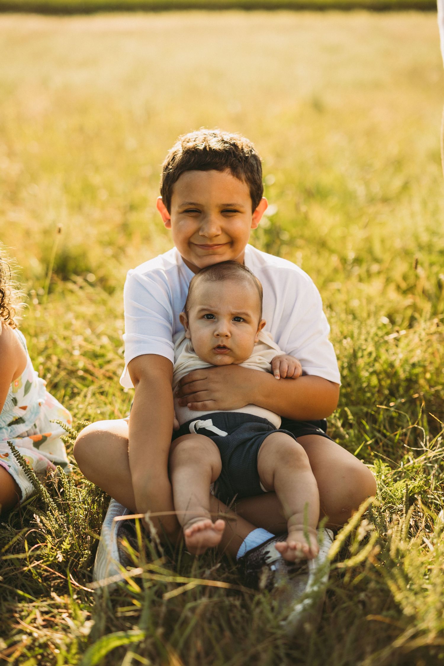 Smiling older boy in a white shirt sits in a sunny grassy field holding a baby in a gray onesie and dark shorts on his lap.