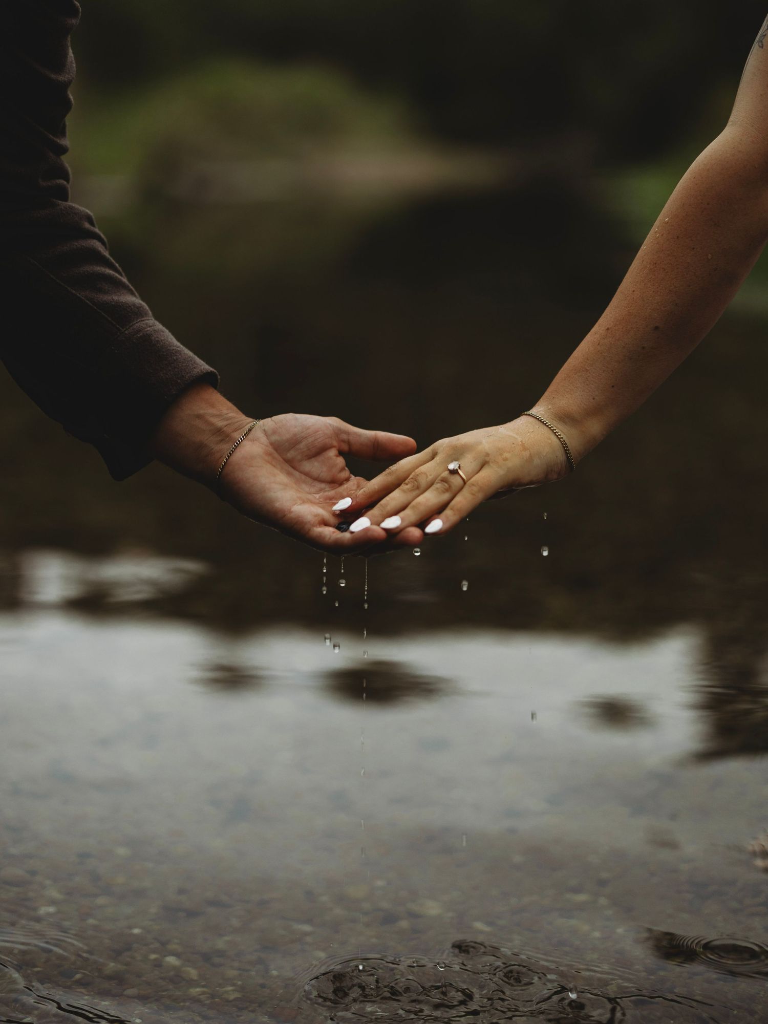 Two hands reaching toward each other over still water, one wearing a ring and bracelet, with droplets falling from their fingertips.