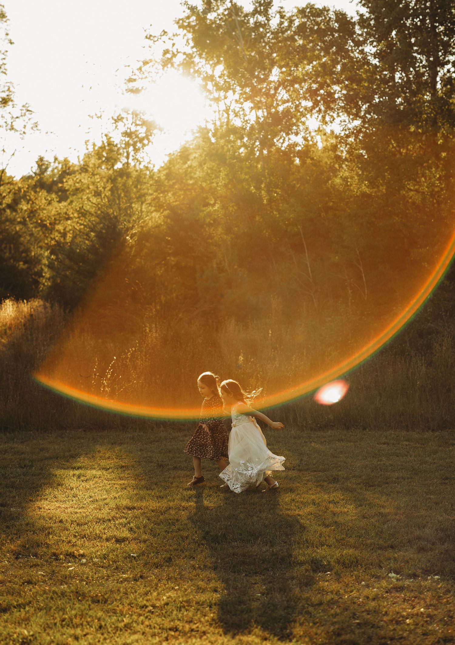 Two children in white and neutral outfits run across a grassy field at golden hour with a glowing orange sun flare behind tall trees.