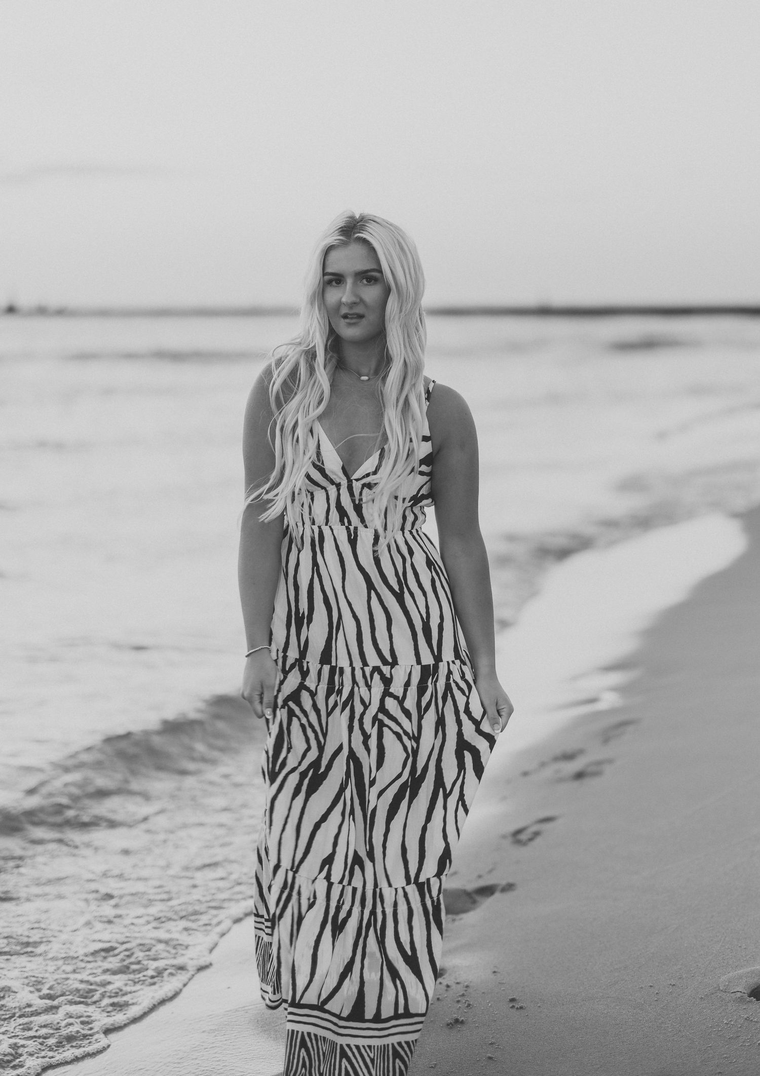 Black and white portrait of a woman in a zebra print maxi dress walking along the shoreline of a beach.