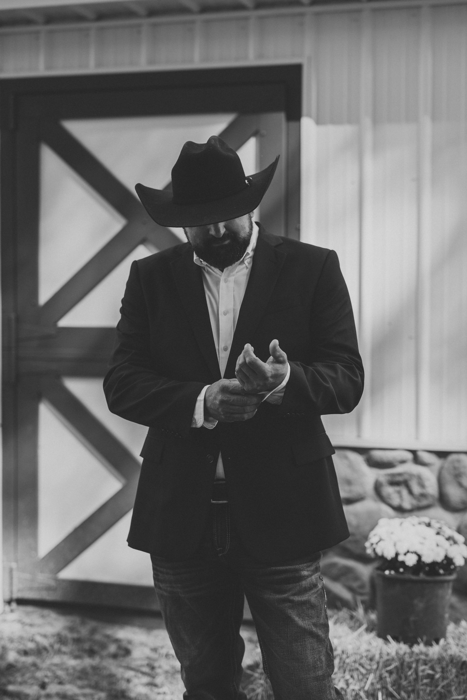 Black and white photo of a bearded man in a cowboy hat and blazer rubbing his hands by a barn door and a pot of flowers.