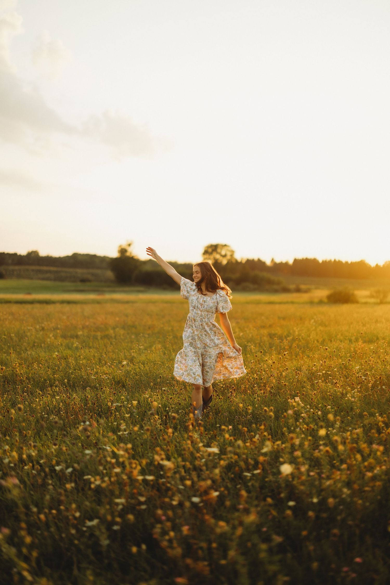 Woman in a floral dress twirls with one arm raised in a golden wildflower meadow at sunset.