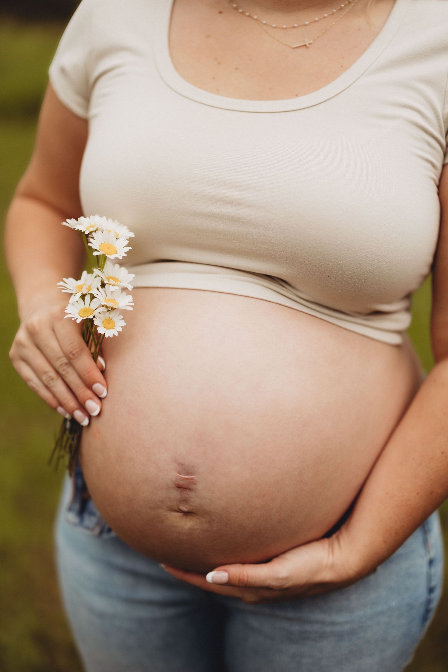 Pregnant woman in beige crop top and jeans holding white daisies while cradling her bare baby bump outdoors.
