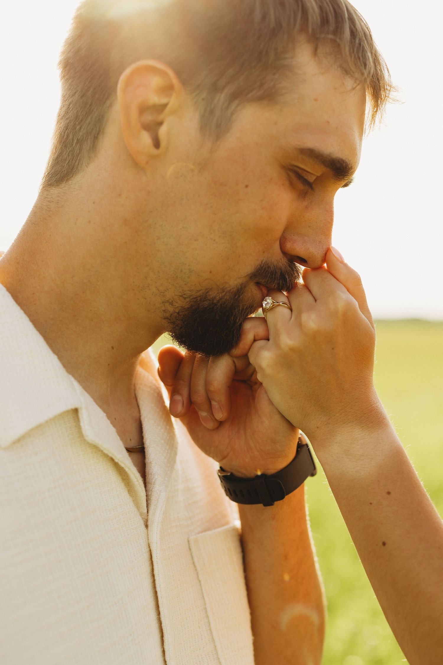 Man with beard kissing woman's hand with engagement ring outdoors in golden sunlight.