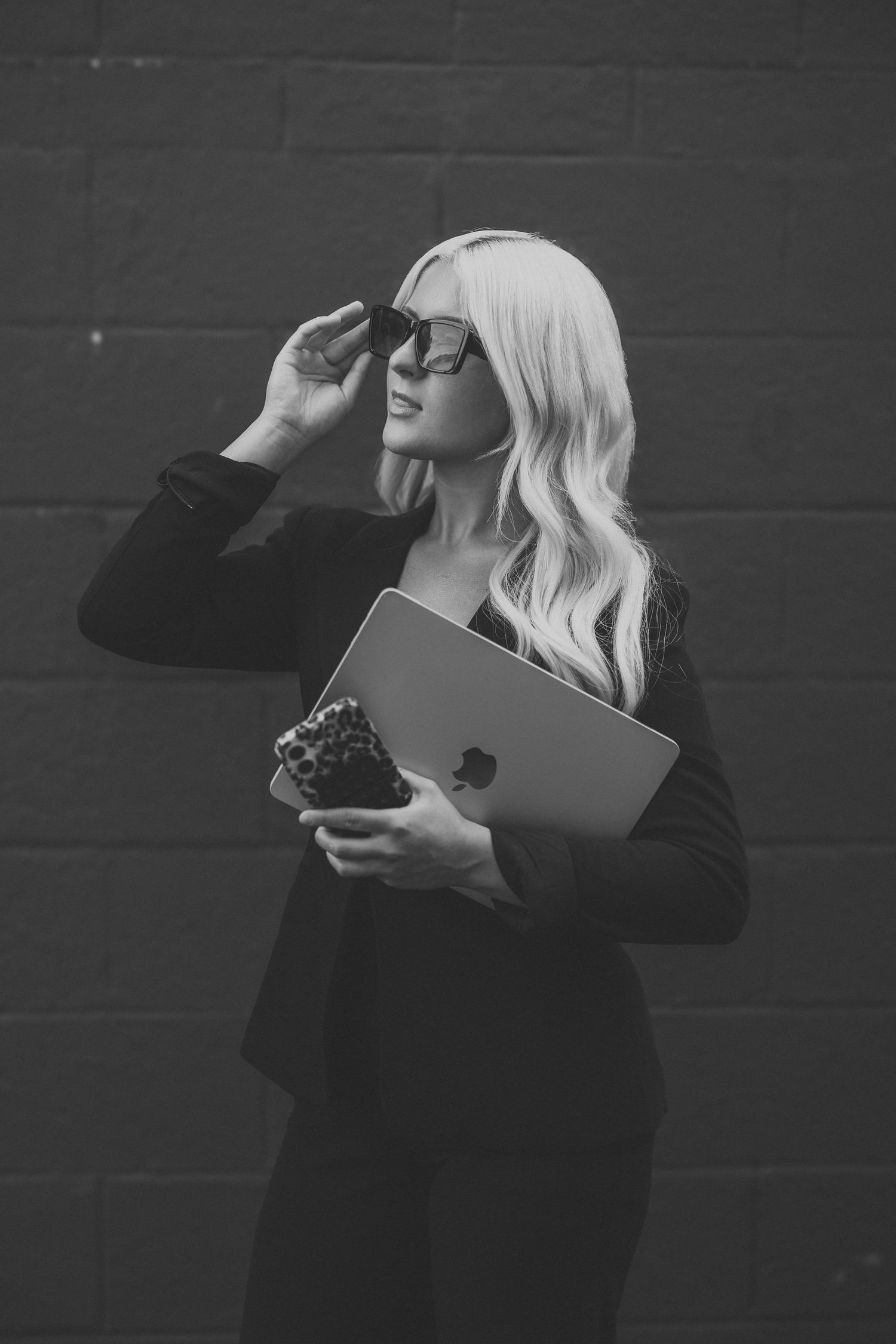 Blonde woman in black outfit holding MacBook laptop and adjusting sunglasses against dark brick wall.