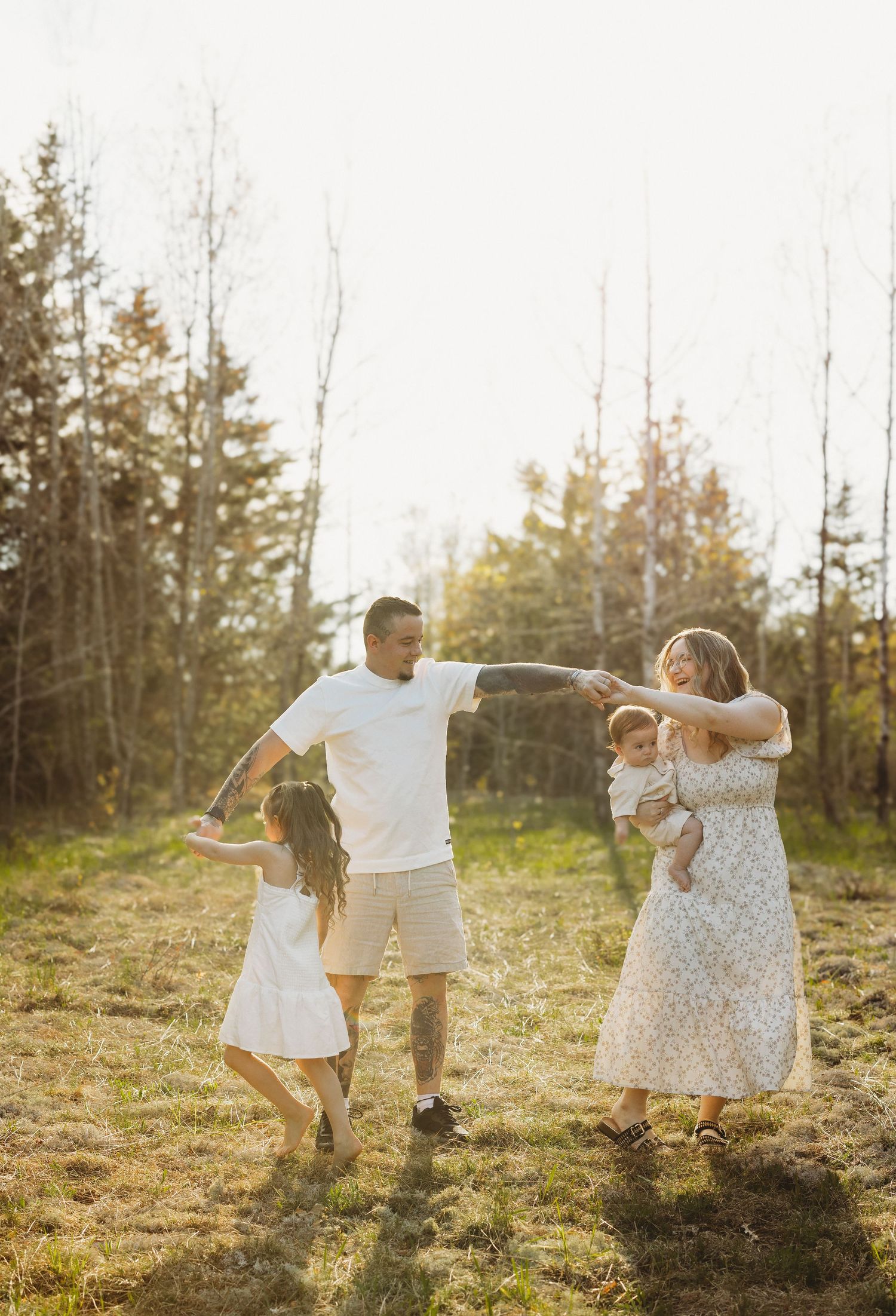 Family of four playing in a sunlit forest meadow, parents and two children in white outfits enjoying a candid outdoor moment.