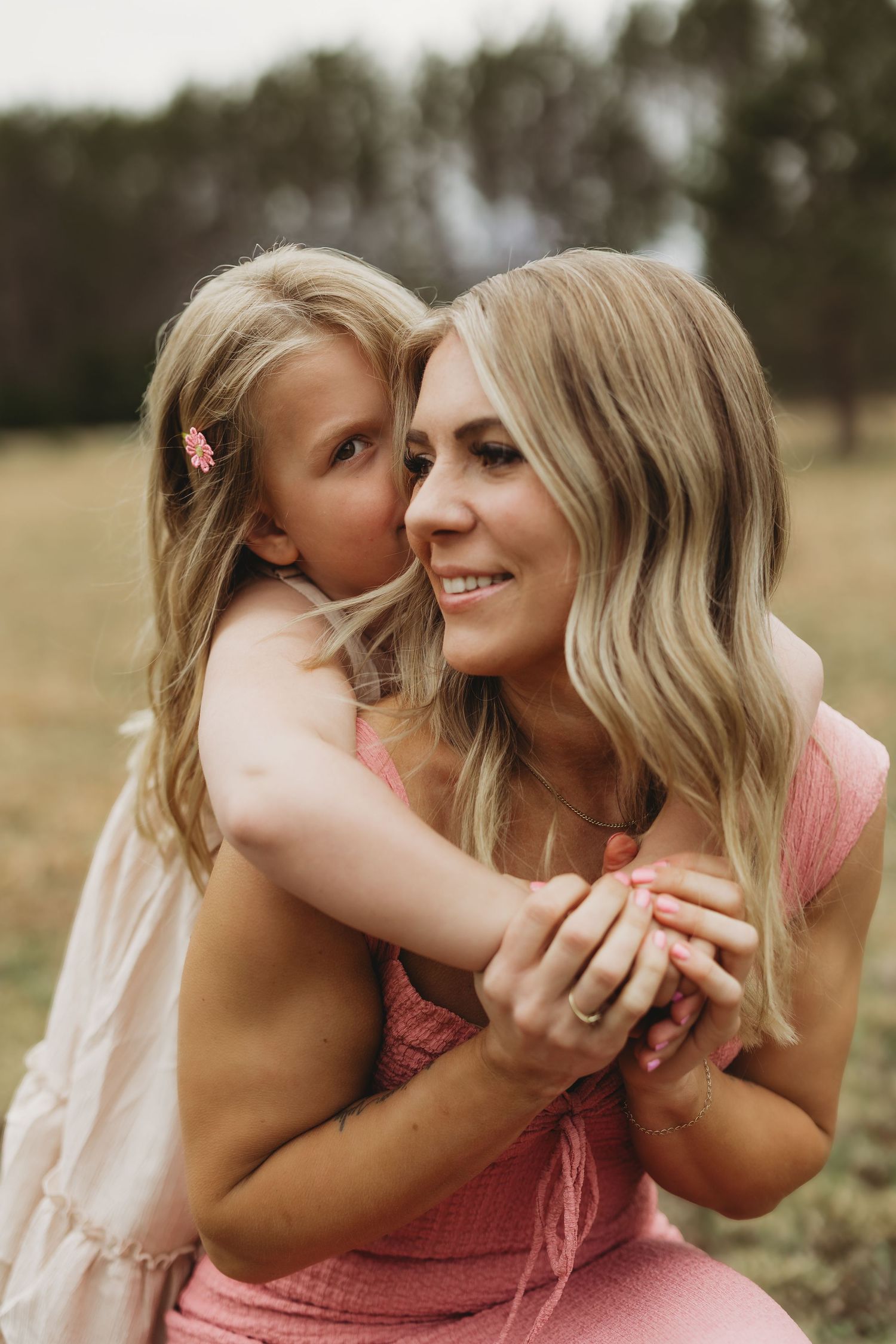 A young girl hugs and whispers to a smiling blonde woman in a pink dress outdoors in a field.