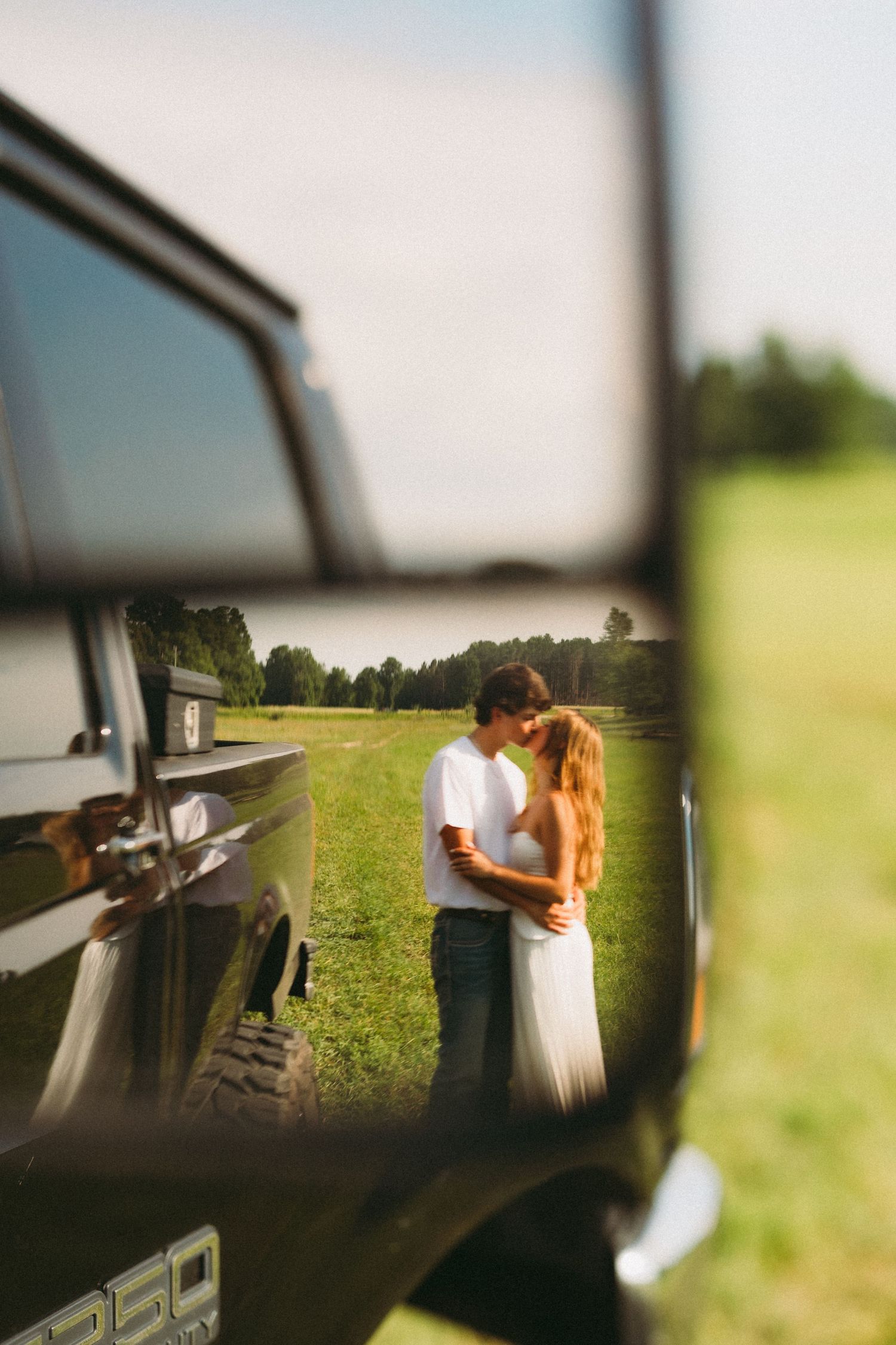 Couple sharing a romantic kiss in a green field, captured through the side mirror of a vintage truck on a sunny day.