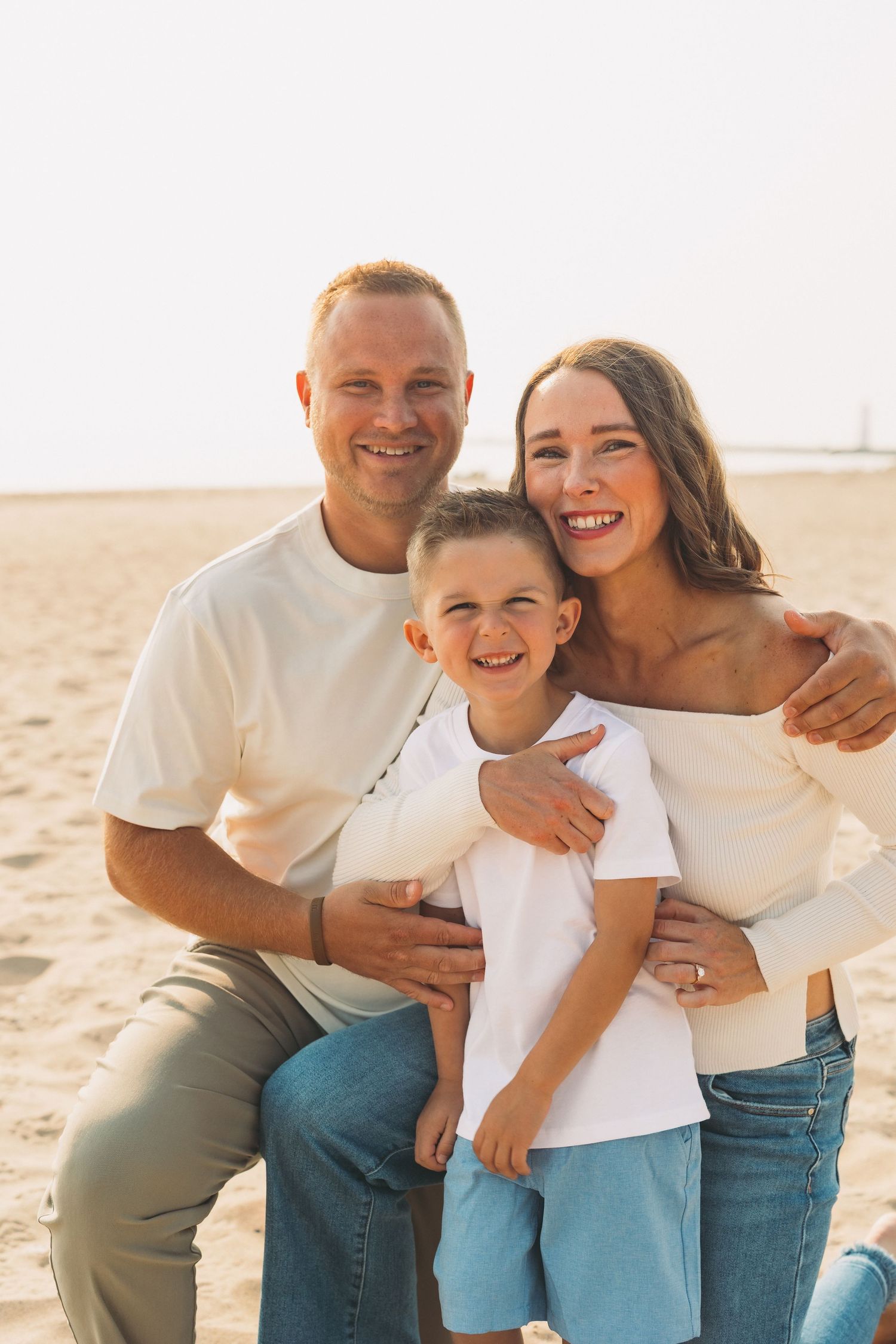 Happy family of three smiling together on a sandy beach, parents embracing young son in matching white outfits.