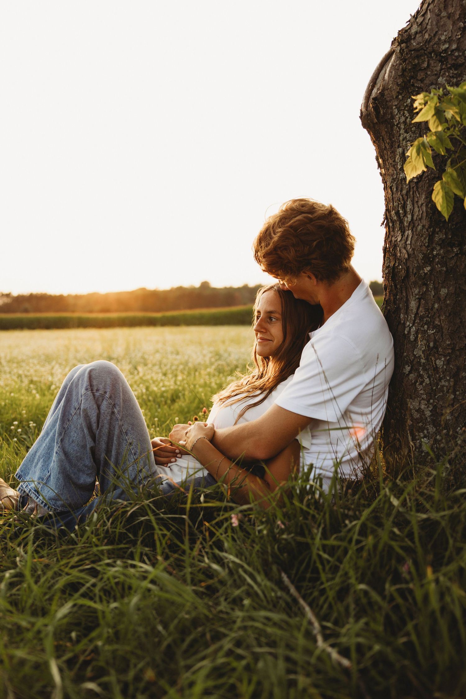 Couple sitting together against a tree in a sunlit field at golden hour, sharing a romantic embrace.