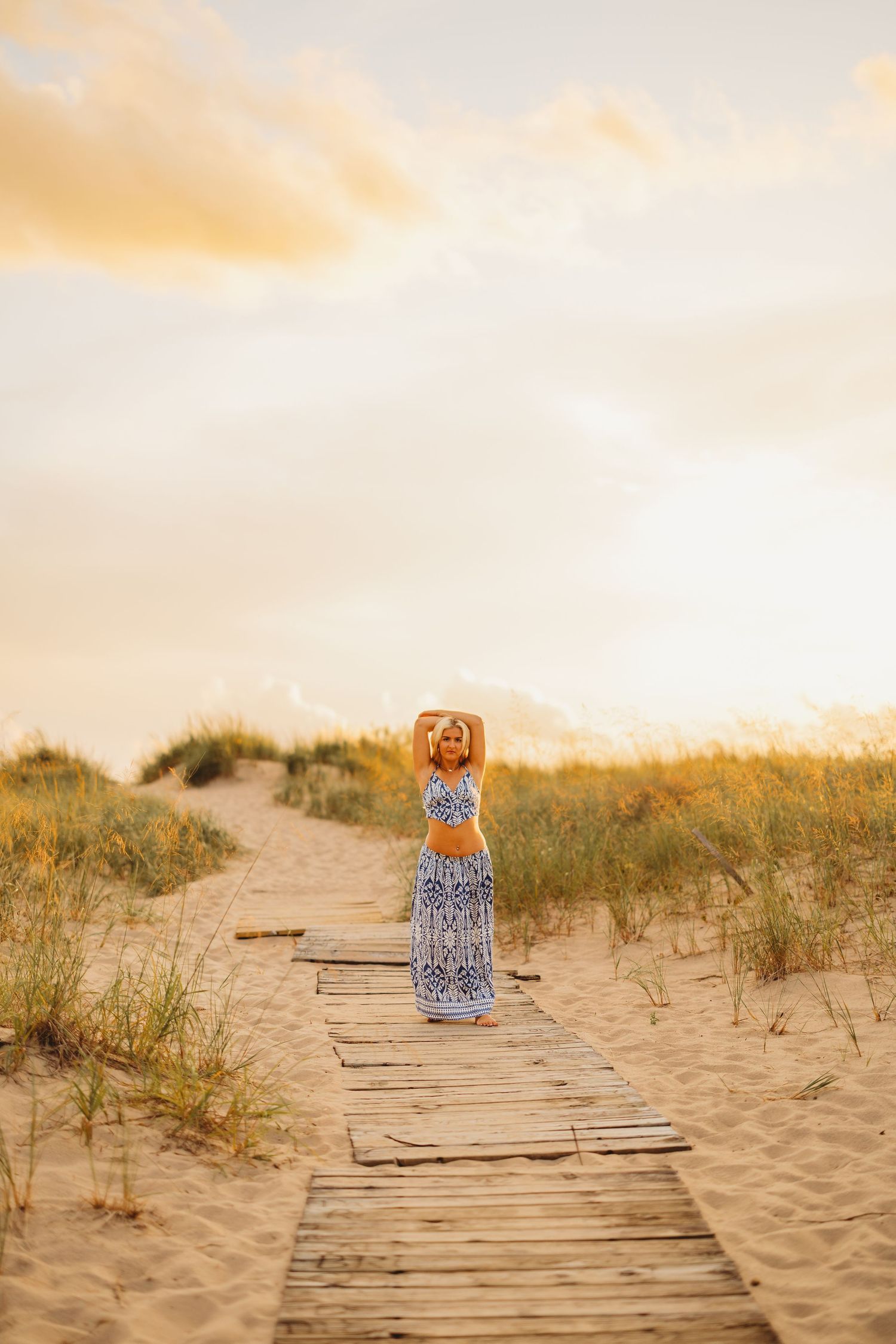 Woman in a blue and white patterned two-piece outfit poses with arms raised on a wooden boardwalk through beach dunes at golden hour sunset.