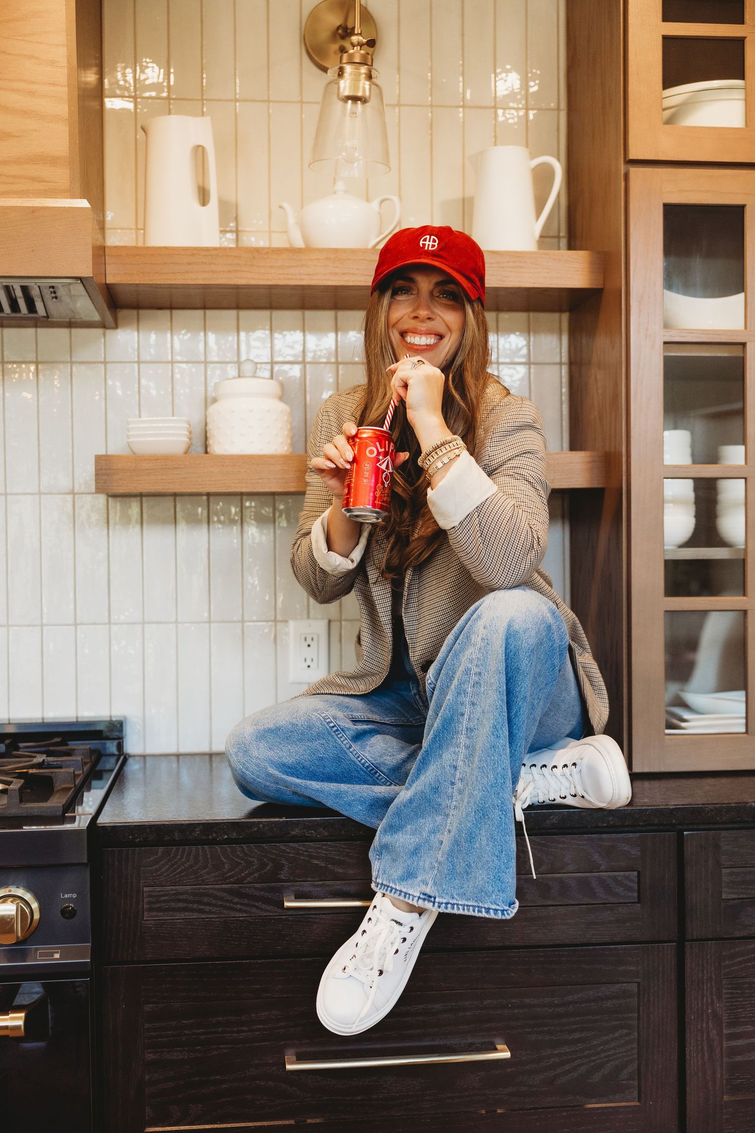 Smiling woman in a red cap, wide-leg jeans and white sneakers sits on a dark kitchen counter sipping a red soda can.