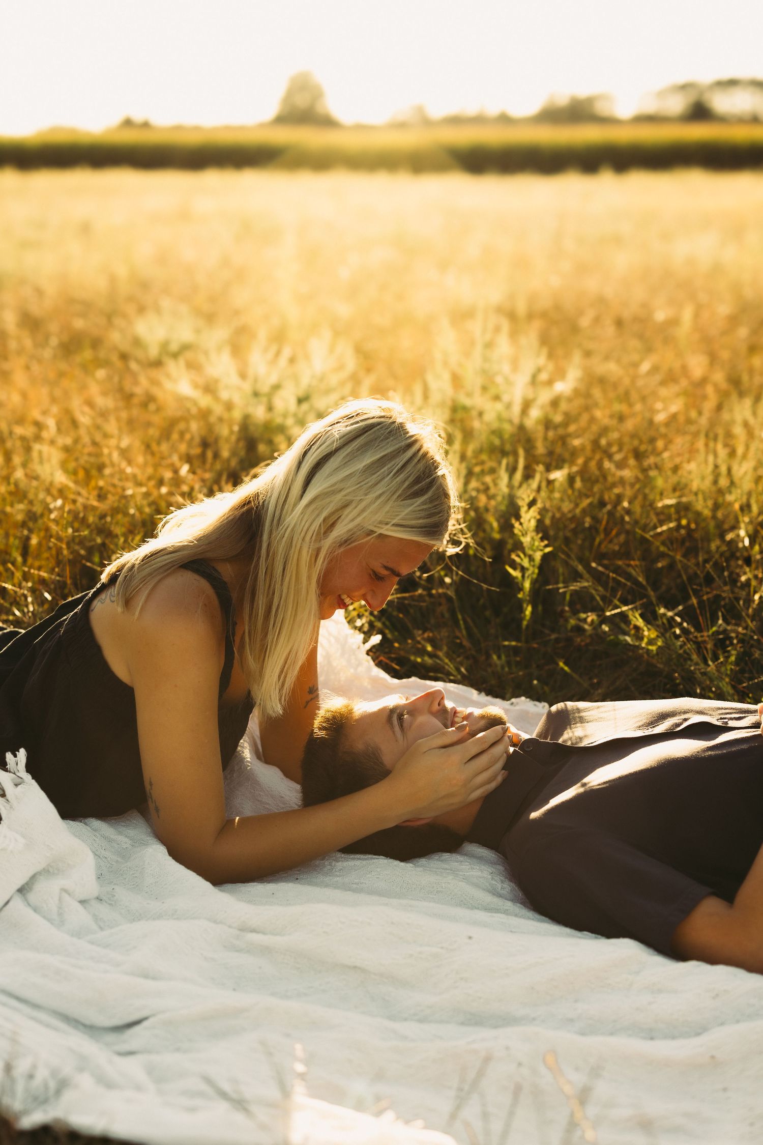 Blonde woman leaning over a person resting on a white blanket in a golden wheat field, glowing in warm sunset light.