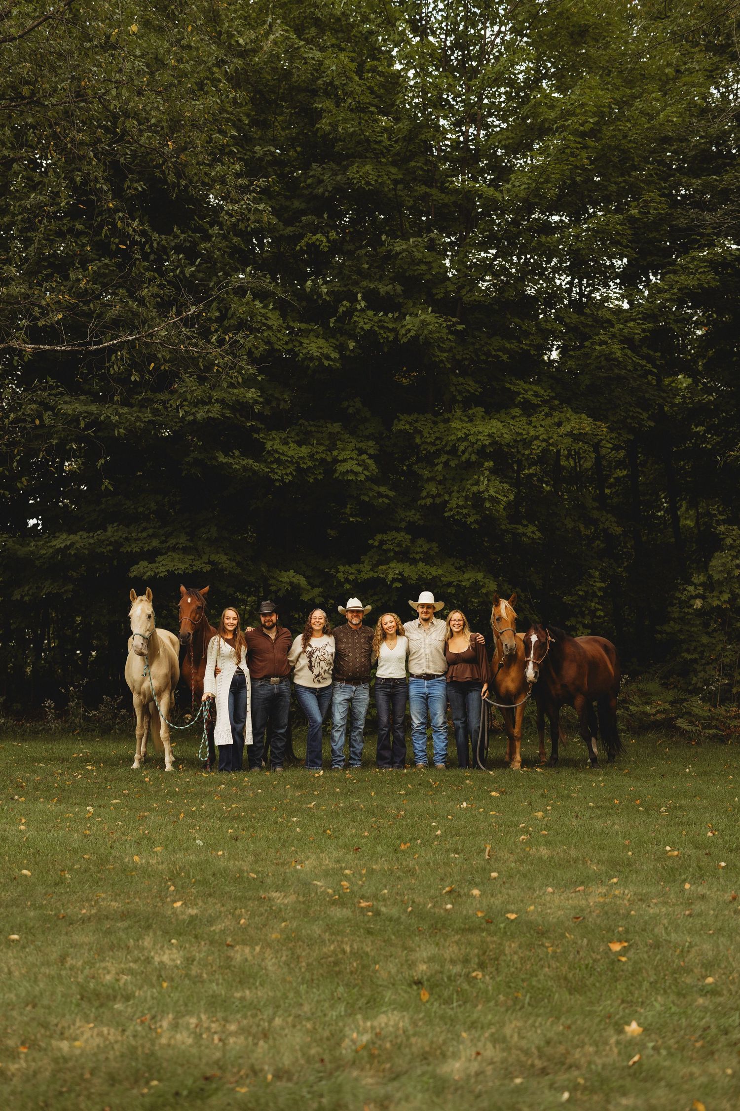 Family of seven in western attire stand arm in arm on a grassy lawn with three horses and tall green trees behind them.