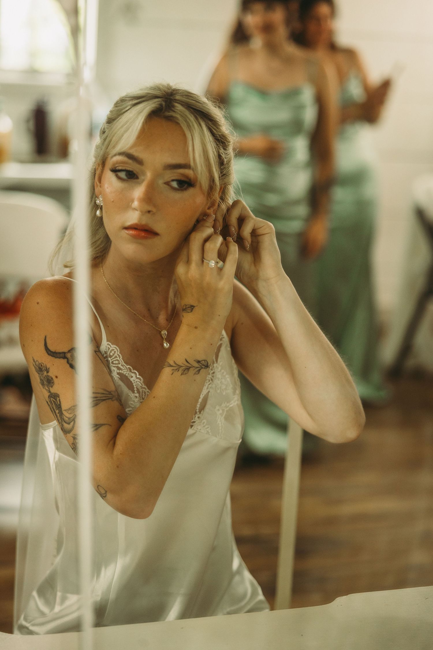 A reflection in a mirror shows a person in a white lace top with visible tattoos and elegant makeup.