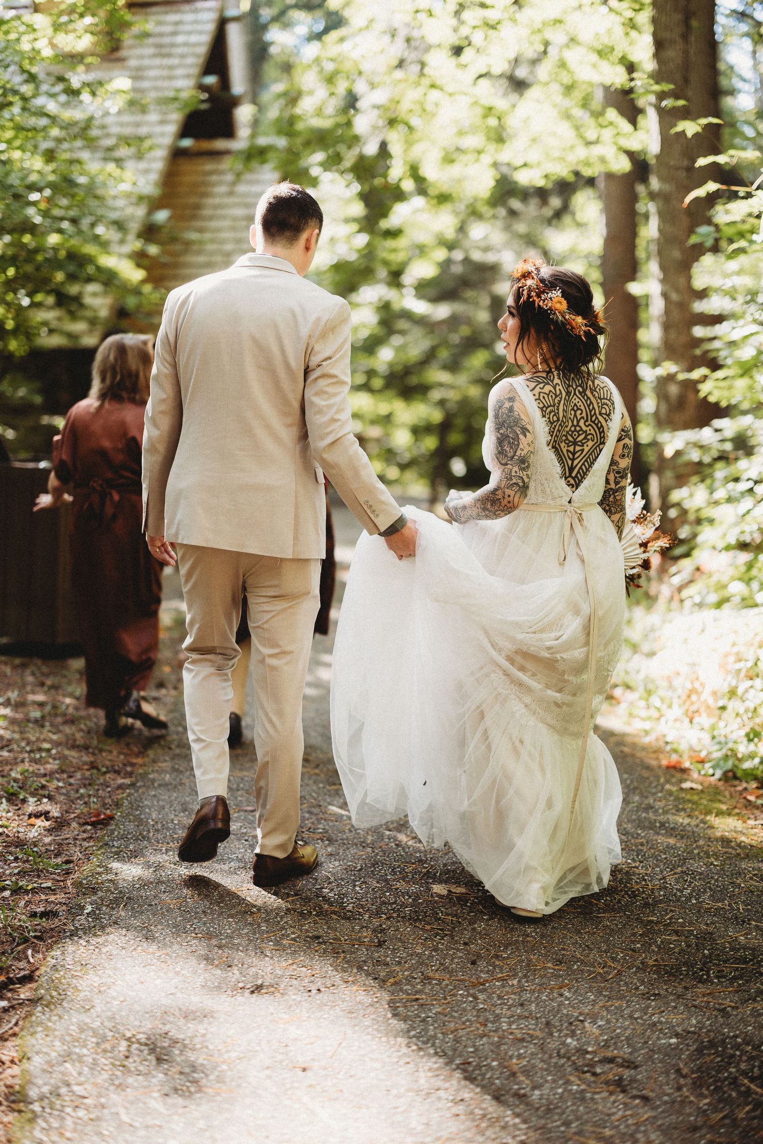 Newlywed couple walks together on a forest path, with bride's lace gown flowing behind them in dappled sunlight.
