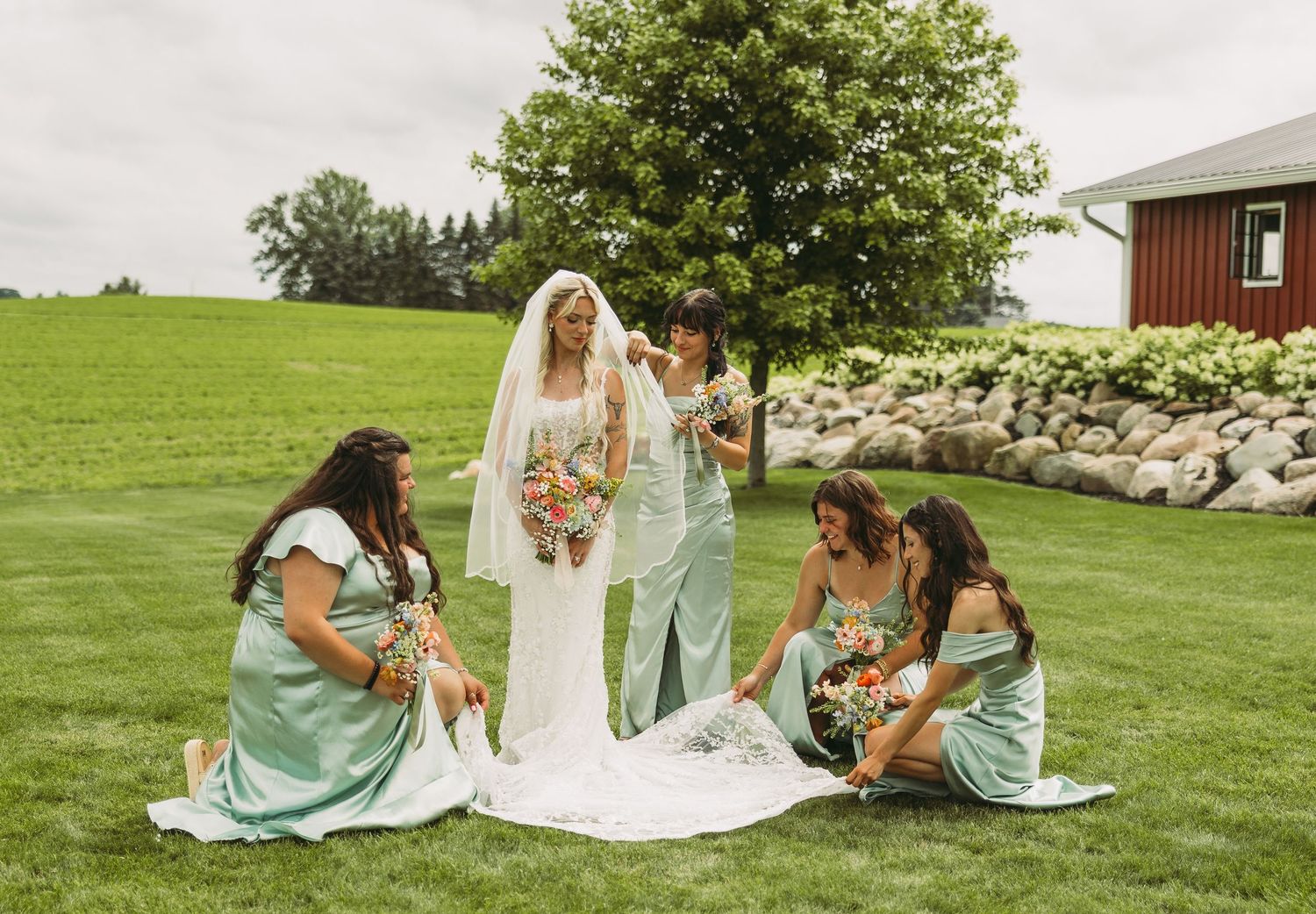 Wedding party relaxes on grass during outdoor photo session near rustic barn.