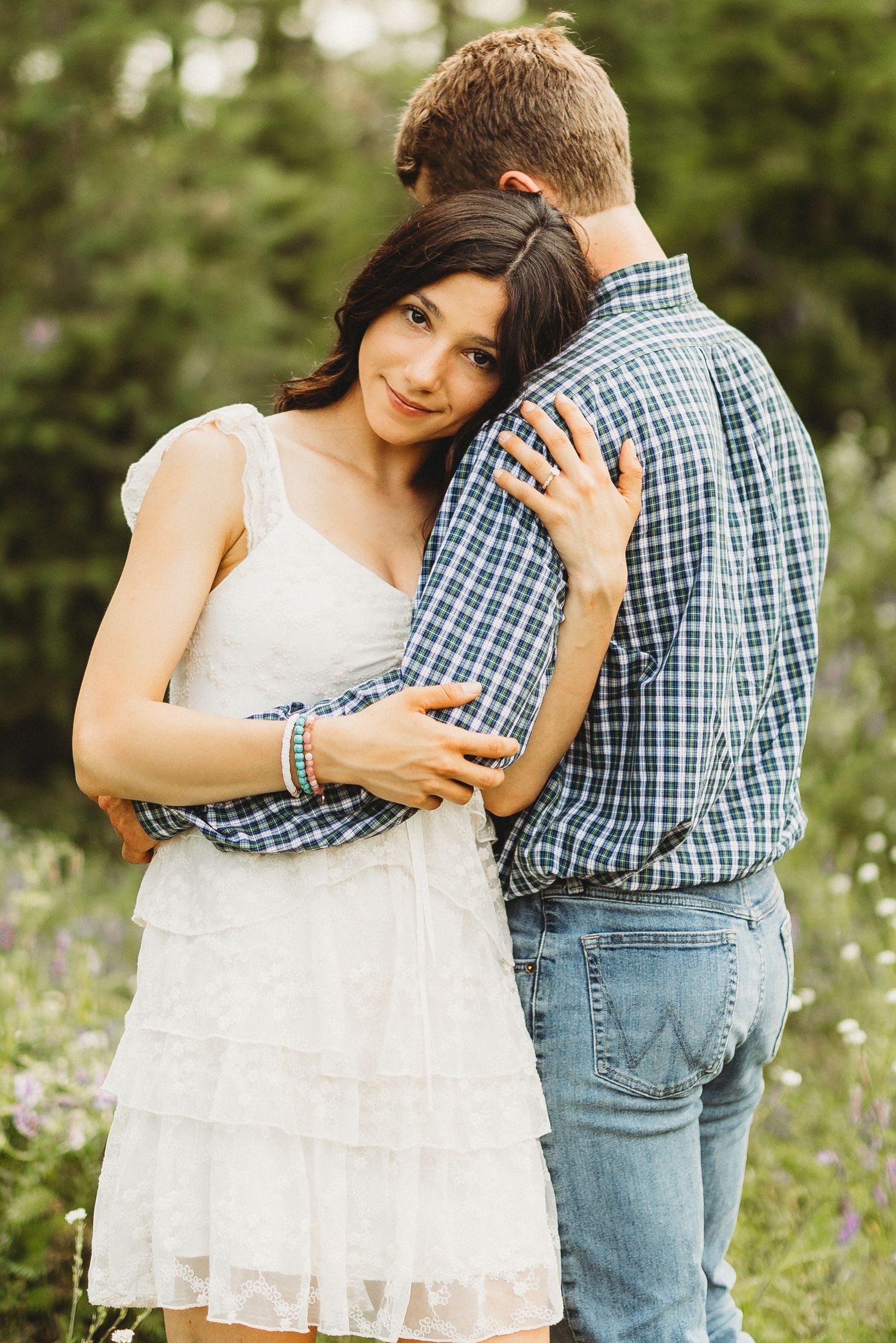 Romantic couple embraces in a wildflower field, with her wearing a white dress and him in a plaid shirt and jeans.