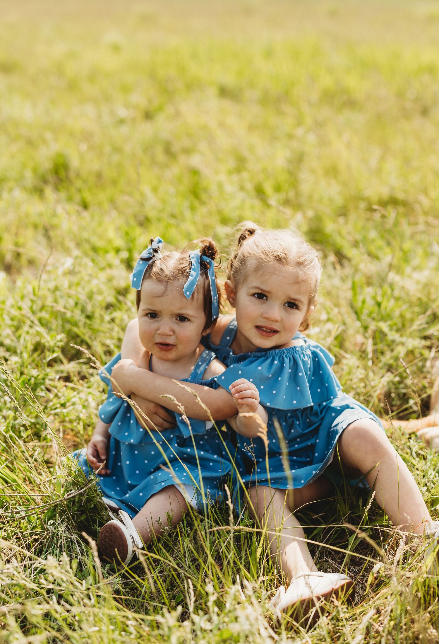 Two young children wearing matching blue outfits lay together in a sunny grass field on a warm day.