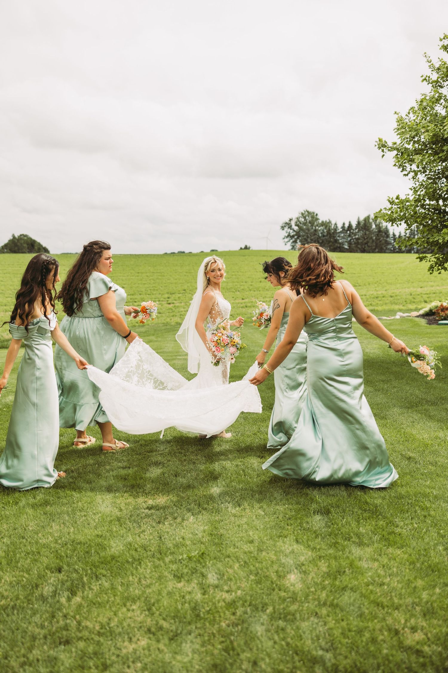 Bridesmaids in mint dresses play with wedding veil on green lawn.