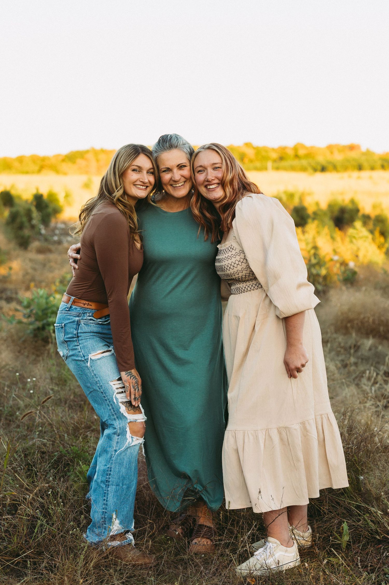 Three women in casual wear hugging and smiling together in sunlit field.
