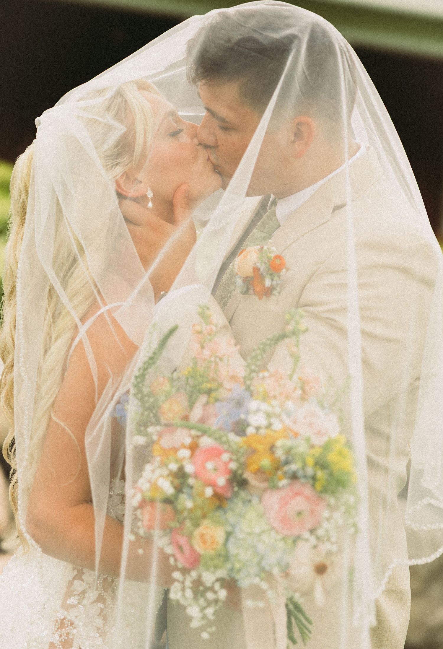 Romantic wedding couple sharing intimate moments under a flowing veil with colorful spring bouquet.