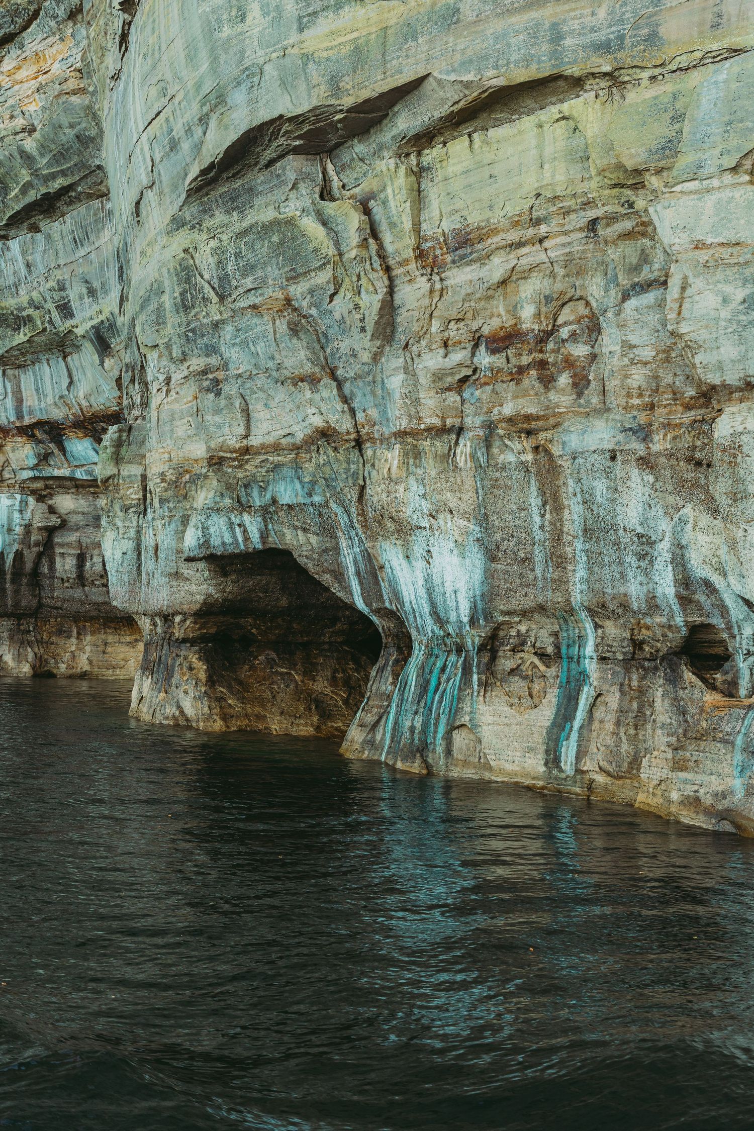 Limestone cliffs with turquoise icicle formations hanging over dark water in a cave system.
