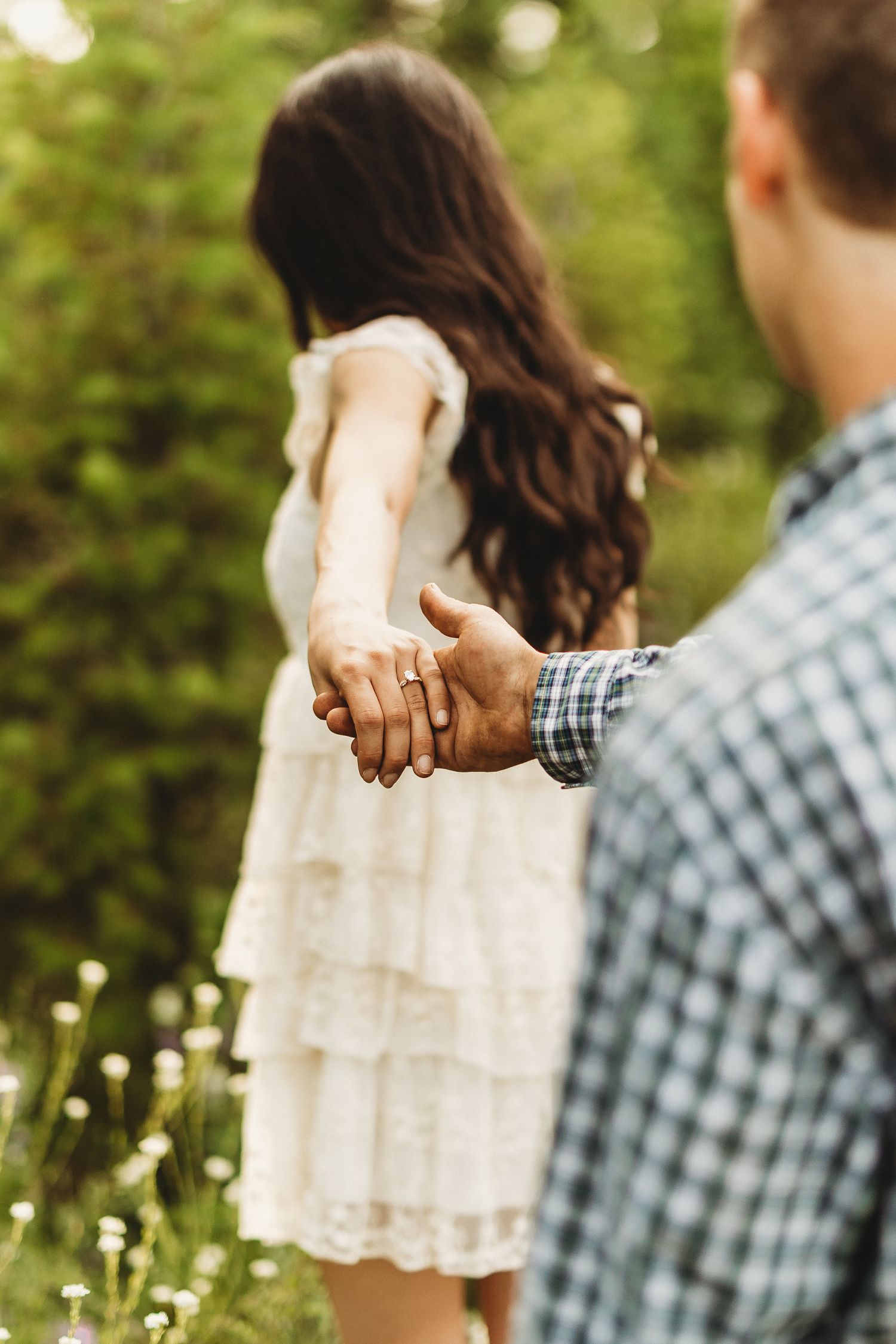 Couple holding hands in a field of dandelions wearing white dress and plaid shirt in soft natural lighting.